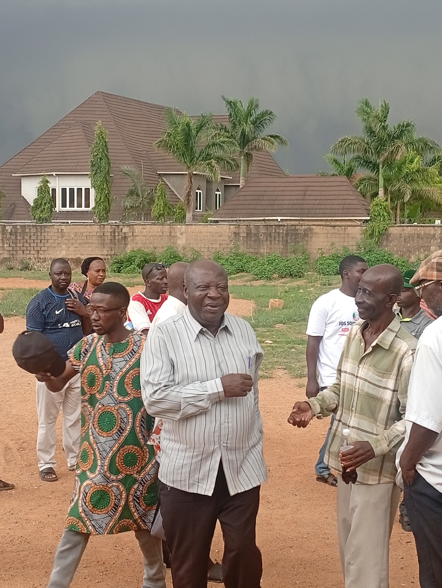 In Bar Jirgi Summa, Nigeria, a group of people stand on a dirt ground. In the background is a tan brick wall and a two-story house with a brown roof, windows, and a dark-colored sky overhead.
A man in a striped shirt is prominently positioned in the center of the group. To the right is a man holding a bottle and wearing a plaid shirt and tan pants. To the left of the man in the striped shirt is a person wearing a patterned shirt. Behind them are other individuals, including one in a blue shirt. Palm trees and green vegetation are visible near the house and wall.