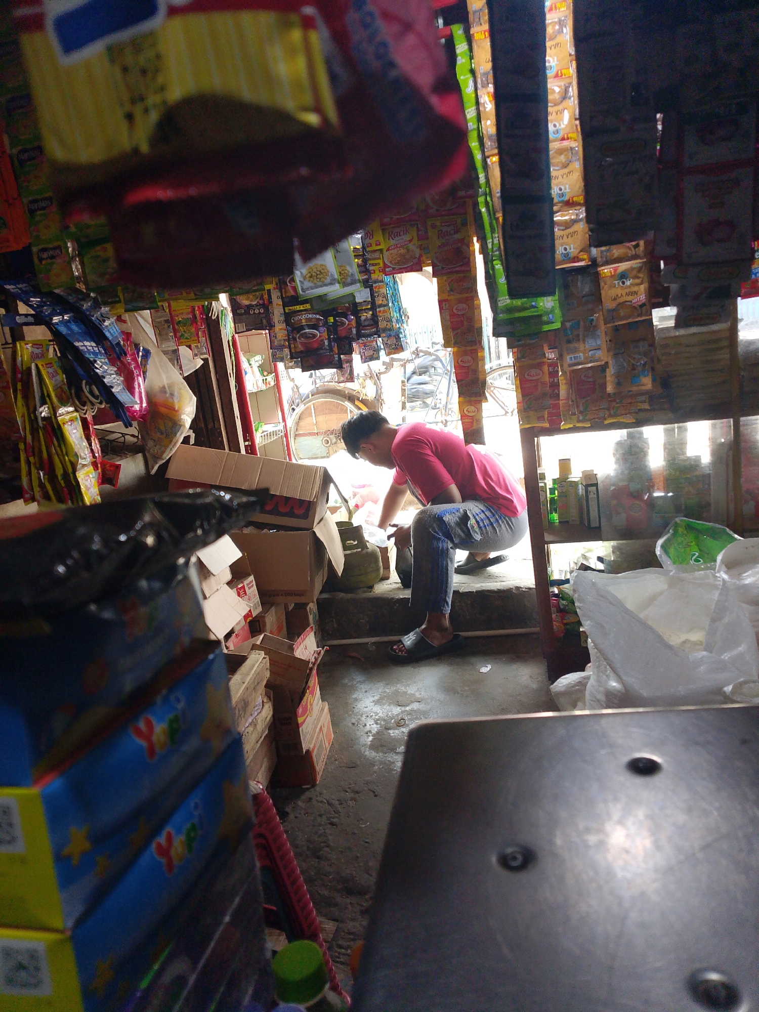 The image depicts the interior of a small convenience store, likely in Palembang, Indonesia, given the context. A young man wearing a pink t-shirt and patterned sweatpants is crouched down, interacting with items on a low step or ledge within the store. The store is packed with merchandise displayed on shelves and hanging from the ceiling. Products visible include various packaged snacks, drinks, and other retail items. Cardboard boxes are stacked around the area where the man is working. Sunlight streams in from an open doorway or window at the back of the store, illuminating dust particles and creating bright highlights on the merchandise. In the foreground, a portion of a metal surface, possibly a counter or table, is visible. The overall scene suggests a busy, slightly cluttered retail environment.