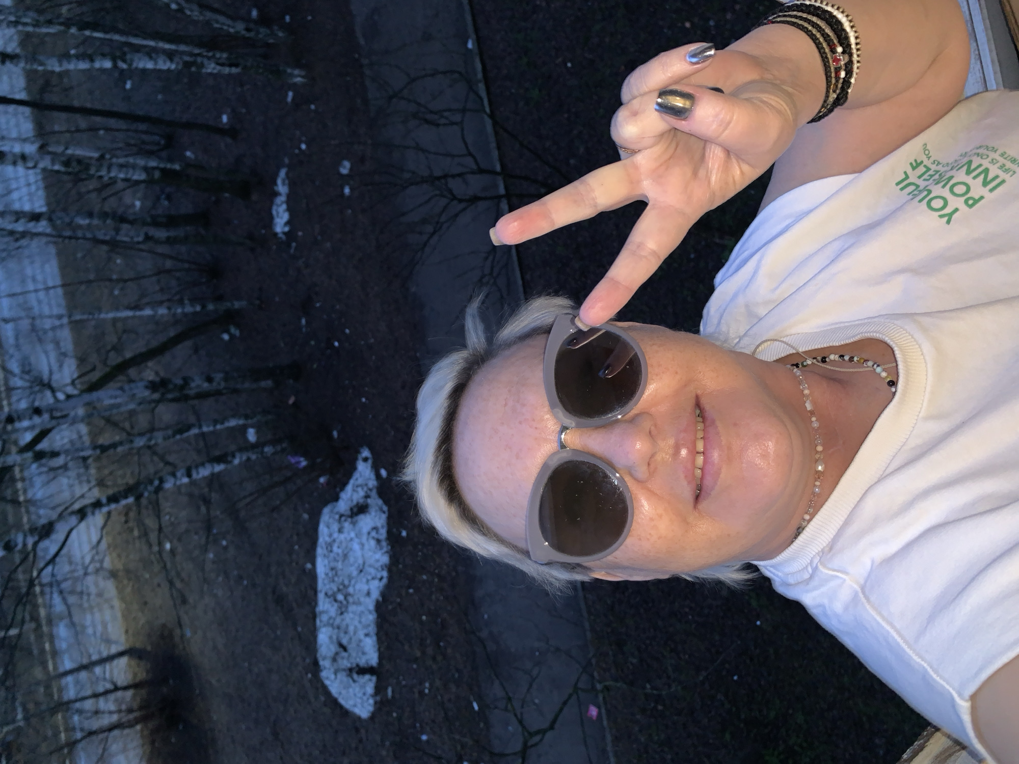 A middle-aged woman with short blonde hair is making a peace sign. She is wearing sunglasses and a white t-shirt with green writing that says "Your Power Inn". She is also wearing a beaded necklace and several bracelets. She is standing outside, and there is a view of trees and snow on the ground behind her. The location is the city of Chaikovsky, Russia.