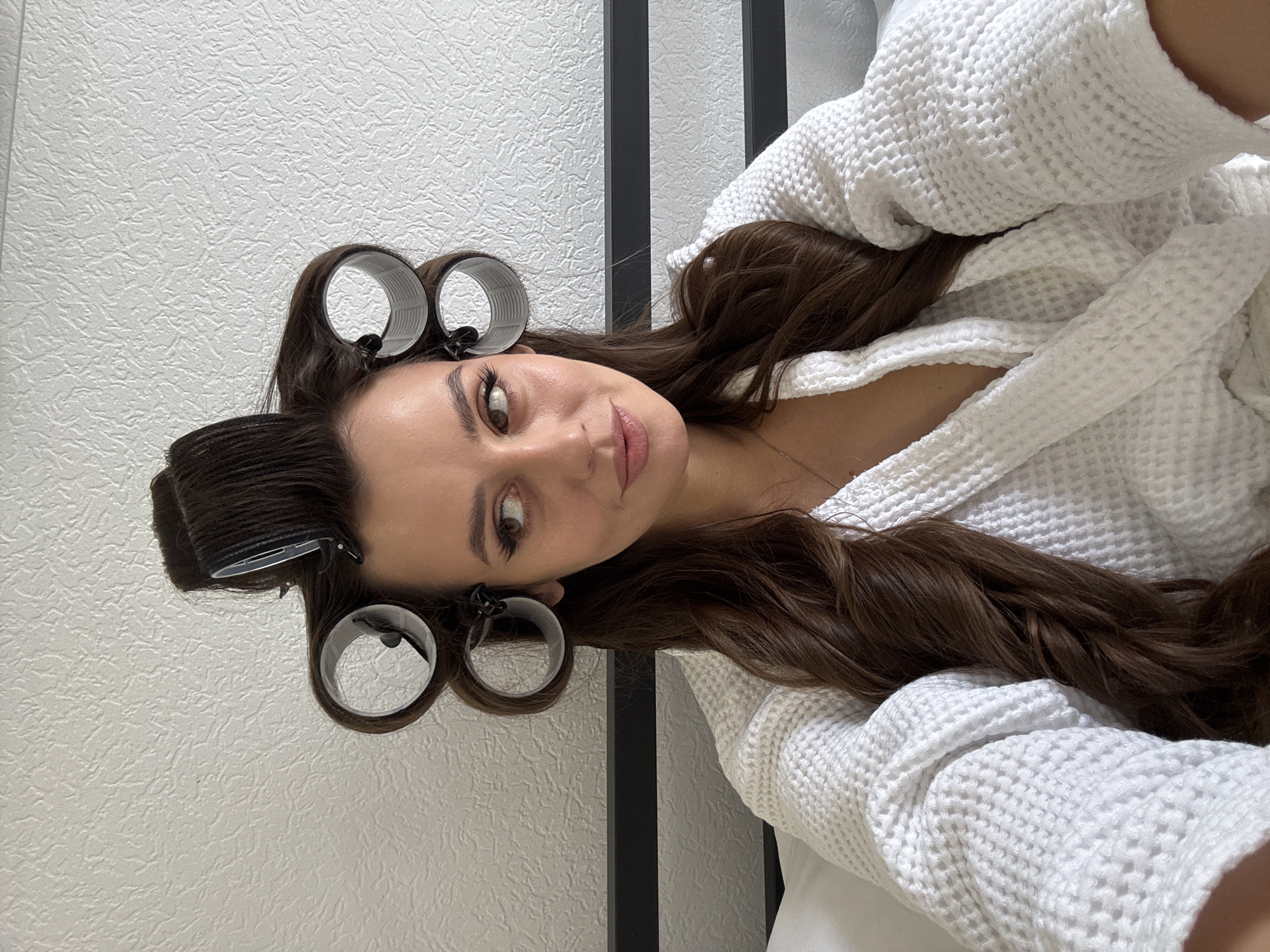 A woman with brown hair is seen wearing curlers and a white bathrobe. She is facing the camera. The background includes a textured white wall and a black metal frame. The location is not identified in the image. The city of Moscow, Russia, cannot be determined.