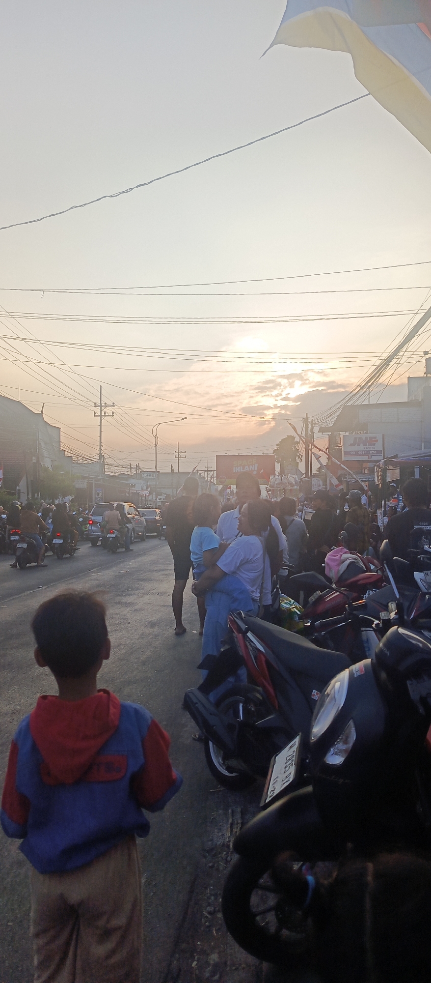 The image depicts a street scene in Malang, Indonesia, bathed in the warm glow of sunset. In the foreground, a young boy faces away from the camera, wearing a blue and red hooded jacket and light brown pants. He stands on a paved street that appears to be busy with traffic. A row of parked motorcycles is visible to the right, with one black scooter prominently featured. In the mid-ground, a group of people are gathered. Among them, a woman holds a child in her arms. The street is lined with buildings, some of which have signs. Power lines crisscross overhead, characteristic of urban Indonesian landscapes. The sky is partly cloudy, with the sun peeking through, creating a hazy and diffused light. Further down the street, vehicles, including cars and motorcycles, are moving, suggesting ongoing activity. The overall impression is of a vibrant, everyday scene in an Indonesian city at dusk.