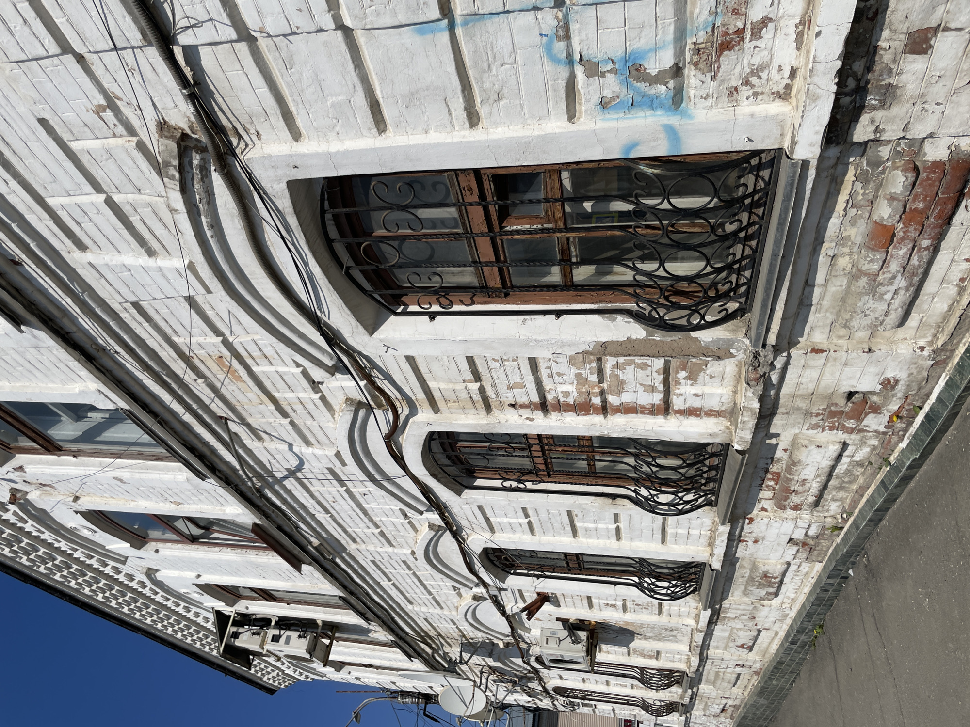 The image depicts the exterior of a building in Izhevsk, Russia. The structure has a white-painted brick facade with decorative elements, including projecting horizontal courses and arched window openings. Each window is protected by a wrought-iron grille with ornate circular designs. Paint is peeling from the brick in some areas. A bundle of electrical cables is affixed to the exterior wall, running vertically down the building's face. The bottom of the image shows other appliances and equipment. The image is taken from a low-angle perspective. The sky is visible at the lower left corner, and a section of a sidewalk or pavement is on the right.