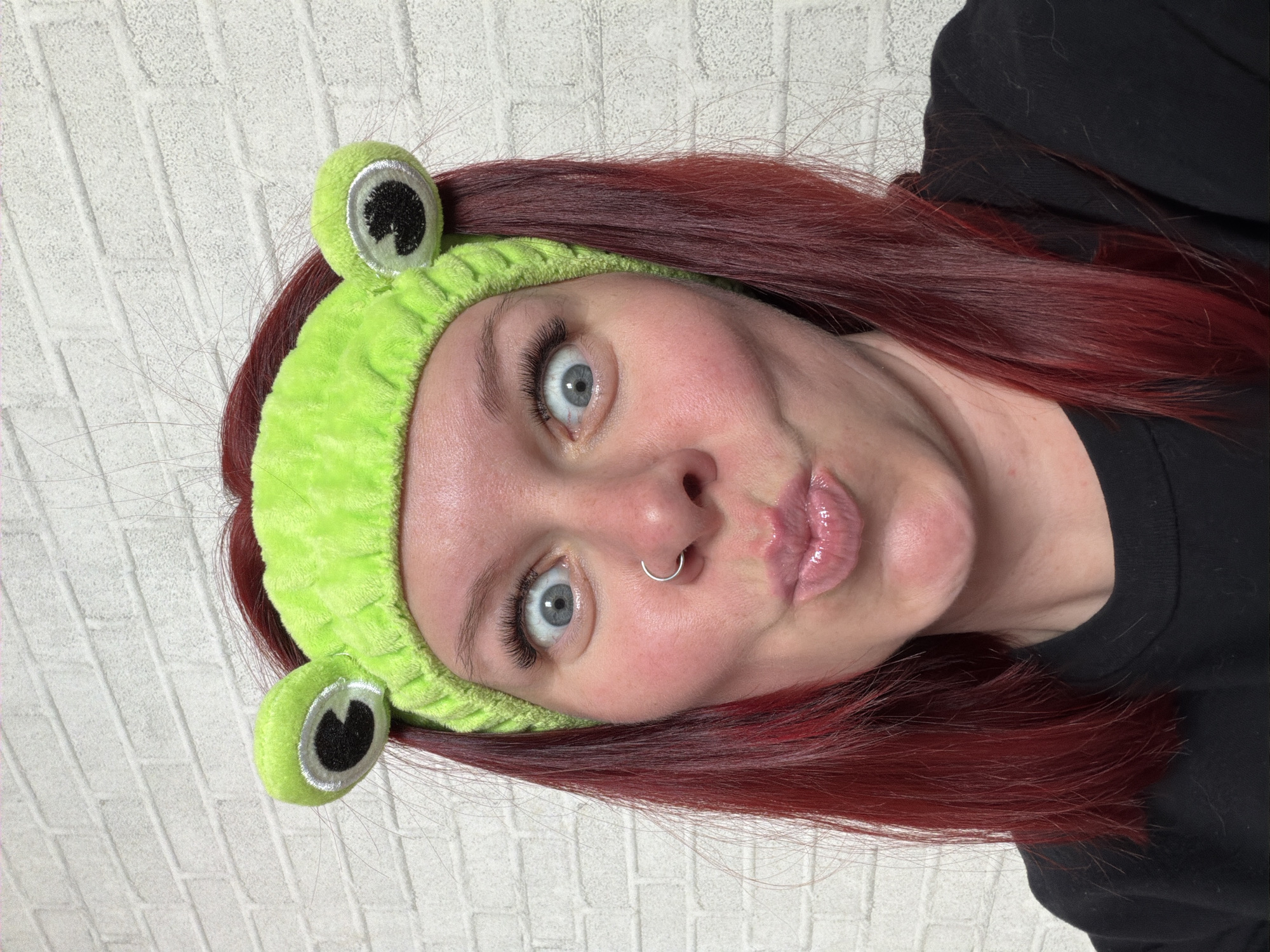 A female individual with red hair and fair skin is depicted in a medium close-up, facing the viewer directly. She wears a bright green, plush headband featuring two prominent, spherical frog-like eyes with black pupils, white sclera, and silver rims, positioned above her forehead. Her light blue eyes are wide open, and her lips are puckered forward in an 'o' shape, appearing glossy. A silver-colored ring pierces her left nostril. She is clothed in a plain black, crew-neck garment, visible from the shoulders up. The background consists of a textured, light-colored wall with a repeating rectangular brick pattern. The location in the city of Chaikovsky, Russia, cannot be determined from the visual information provided in the image.
