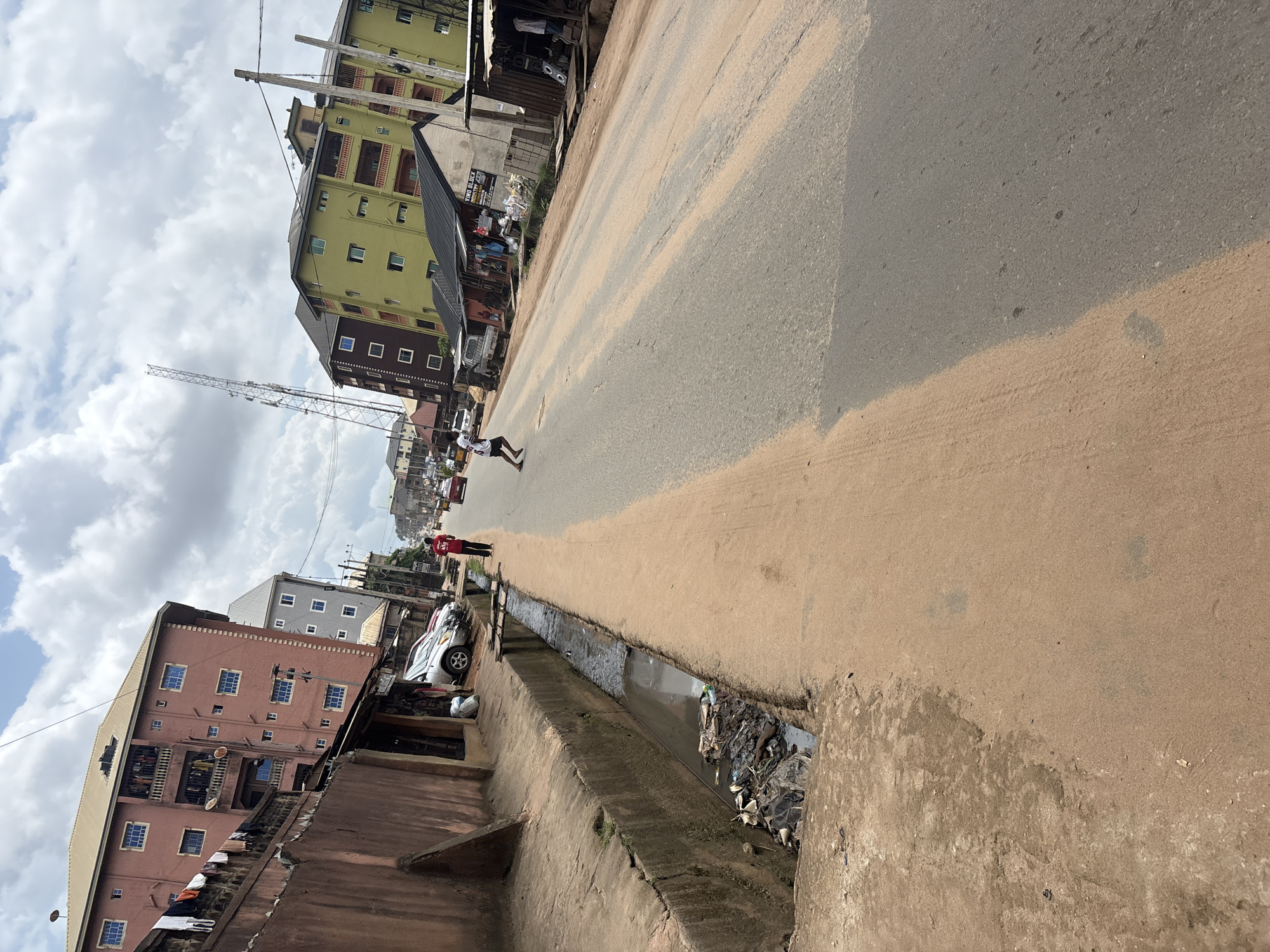 The image depicts a street scene in Nkpor, Nigeria. The foreground is dominated by a sandy roadside and a drainage ditch filled with debris. A paved road stretches into the distance, lined with buildings of various heights and colors, including a pink, a grey, and a lime green multi-story structure. Several cars are parked along the side of the road. Two individuals are visible on the road: one wearing a red shirt and black pants, standing near the drainage ditch, and another person, possibly a child, in a white shirt and black shorts, appearing to jump or run on the road. The sky is partly cloudy. The overall scene suggests an urban environment with evidence of infrastructure and pedestrian activity.