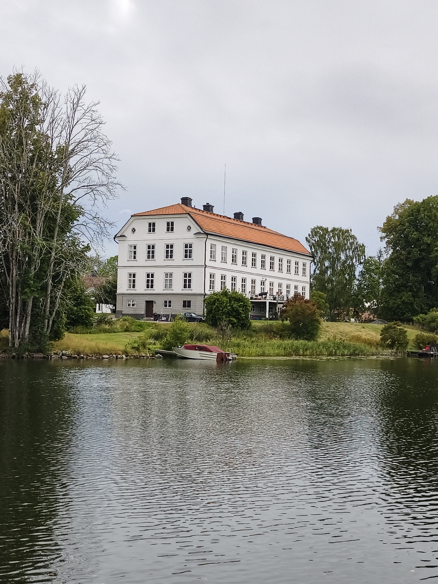 The image shows a large, white building with a red tile roof, situated on the shore of a body of water. The building has multiple windows and appears to be a manor house or a similar grand residence. To the left of the building are several tall trees. In the water, a small boat with a red cover is moored to a dock. The water is calm with ripples reflecting the overcast sky. The background consists of more trees and a grassy slope leading up to the building. The scene is located in Brokind, Sweden, based on the provided context. There are no people visible in the image.