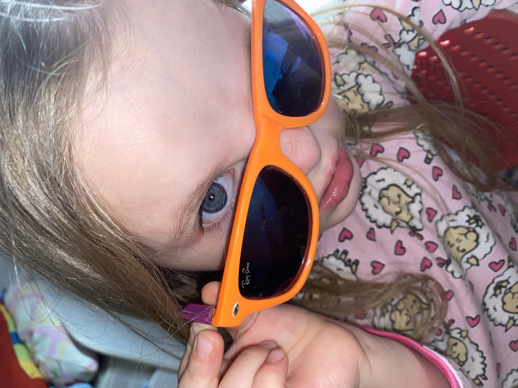 A young child is posing for a selfie while holding a pair of orange-framed Ray-Ban sunglasses. The child has fair skin, light brown hair, and blue eyes. The child is wearing a pink pajama top with a repeating pattern of sheep and hearts. The location of the photo is unspecified, and the claim it was taken in Votkinsk, Russia, is unsubstantiated without further evidence.