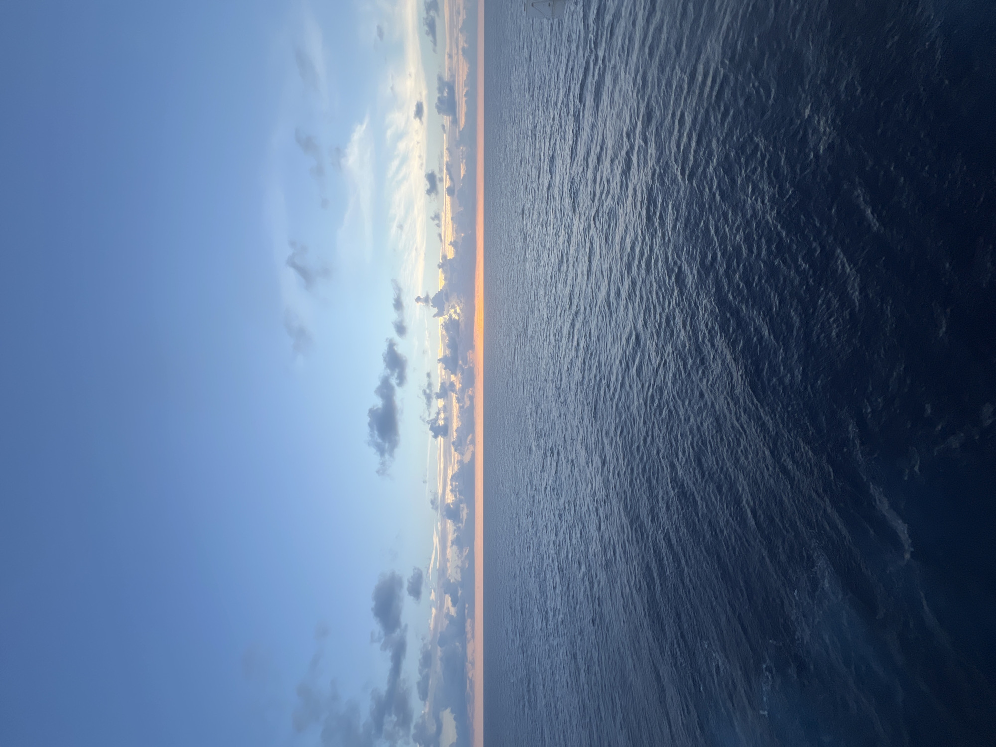 The image depicts a vast expanse of the ocean under a twilight sky. The horizon is a thin, bright line of orange where the sun has set, casting a warm glow that gradually fades into the cooler blues of the sky above. Scattered clouds, some dark and heavy, others wispy and tinged with the sunset colors, dot the sky. The ocean itself is a deep, rich blue, with gentle ripples creating a textured surface. The overall impression is one of peaceful solitude and the immense beauty of nature. The scene is set at an unspecified location, far from any city.