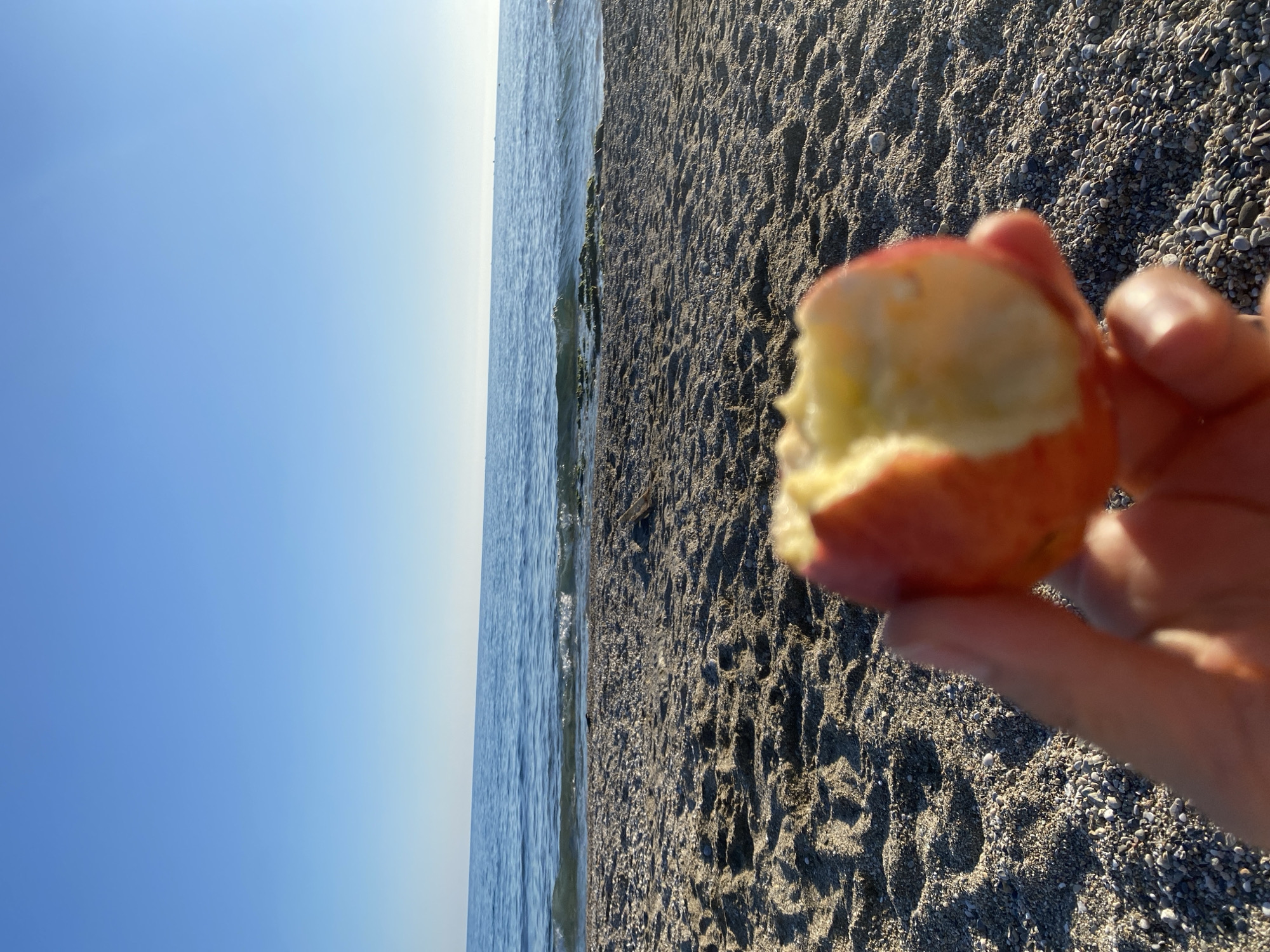 A first-person, eye-level view captures a partly eaten apple held by a person's fingers. The background reveals a beach with grey sand and pebbles adjacent to a calm, blue sea that meets a clear, light blue sky. The scene appears to be at a beach in Alanya, Turkey.