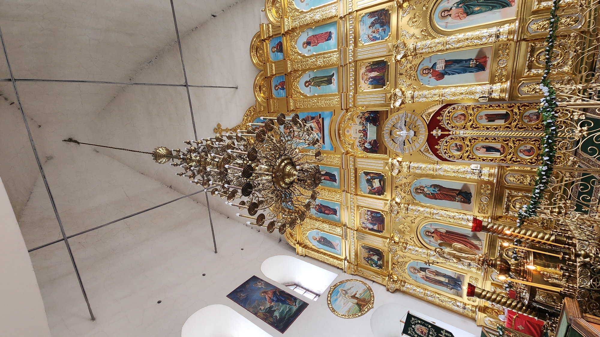 Captured from a low angle looking upwards, the image displays the elaborate interior of a church. A sizable, ornate chandelier dominates the scene, suspended from the ceiling. Below the chandelier, a wall adorned with numerous religious paintings and gold ornamentation is visible. The overall color scheme is rich and warm, with a prominence of gold tones. The architecture seems to be of a historical, possibly Orthodox Christian, design. Natural light streams in through arched windows, illuminating various artworks and decor. The location appears to be within a religious institution, likely in a city of Unknown, Unknown.