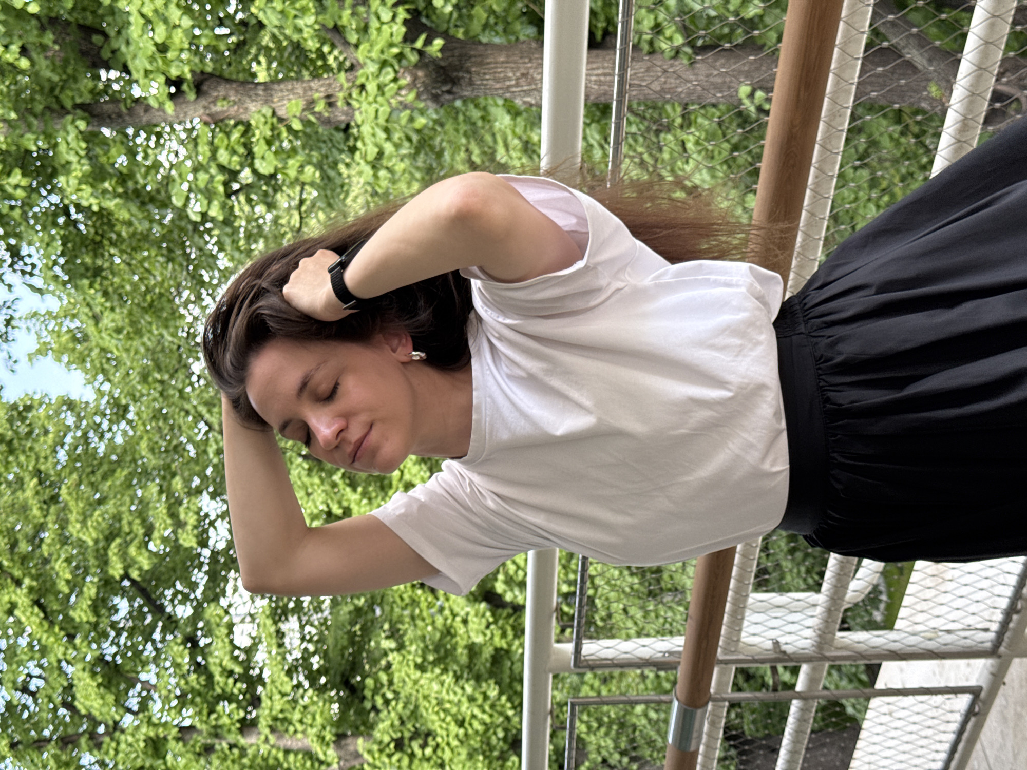 A woman with long brown hair, wearing a white t-shirt and black skirt, stands outdoors, likely on a balcony or elevated platform. She has her eyes closed and her right hand resting on her head. Her left hand is also raised to her head, with a watch visible on her wrist.
The background features lush green trees and foliage, possibly a park or garden. A railing with wooden and metal elements is visible.
The scene seems to be in an urban area, but the specific location in Moscow, Russia, cannot be determined from this image alone.