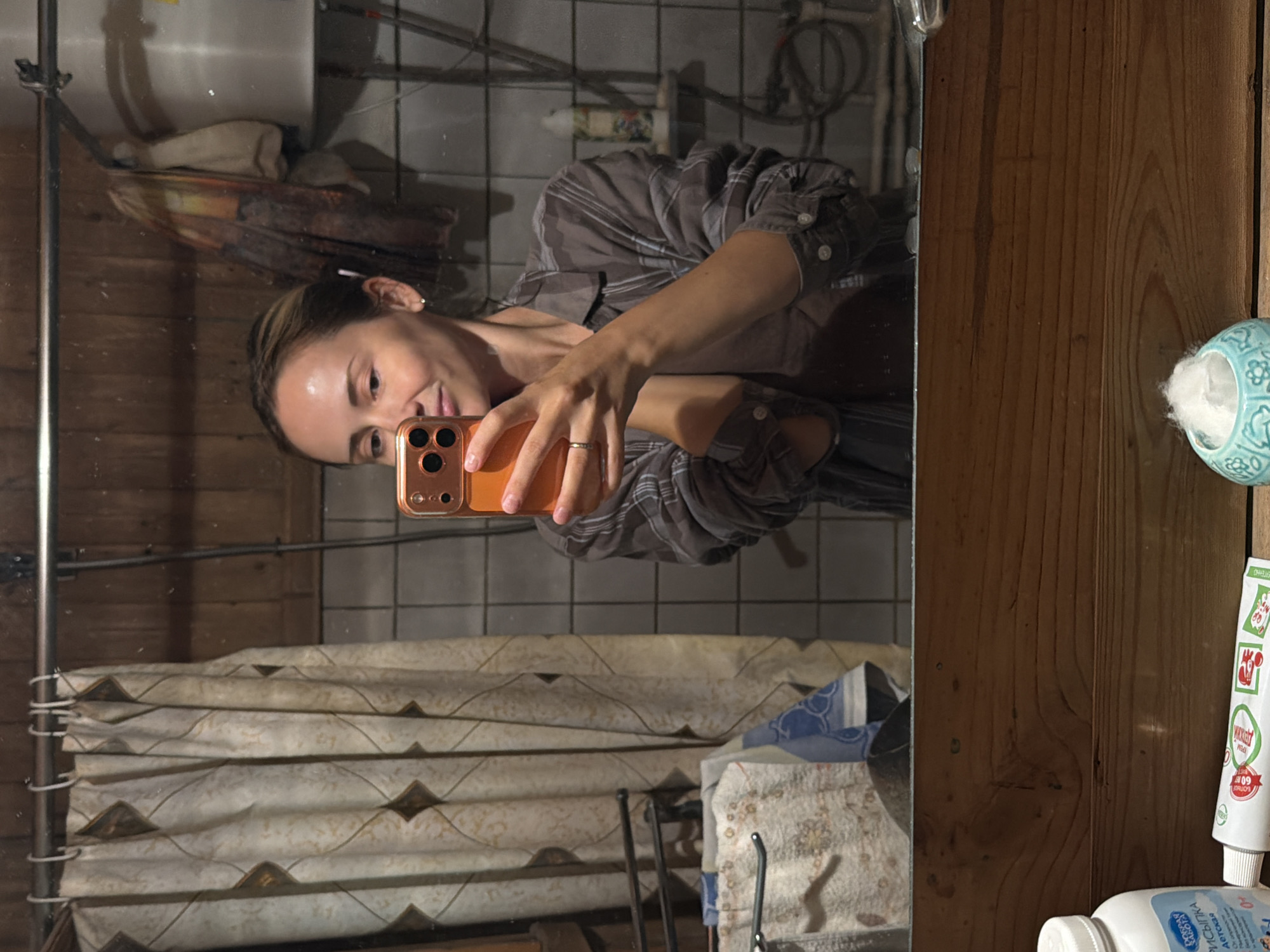A woman with blonde hair pulled back into a bun is taking a selfie in a dimly lit bathroom with a mirror. She is wearing a plaid shirt and a ring on her left hand. The bathroom has tiled walls and a wooden ceiling. A patterned shower curtain is visible on the left, and a shelf with toiletries is on the right. In the foreground, a wooden shelf holds a bottle of baby powder, a tube of toothpaste, and a small teal dish with cotton balls. The image appears to have been taken in a residential setting in Udelnaya, Russia.
