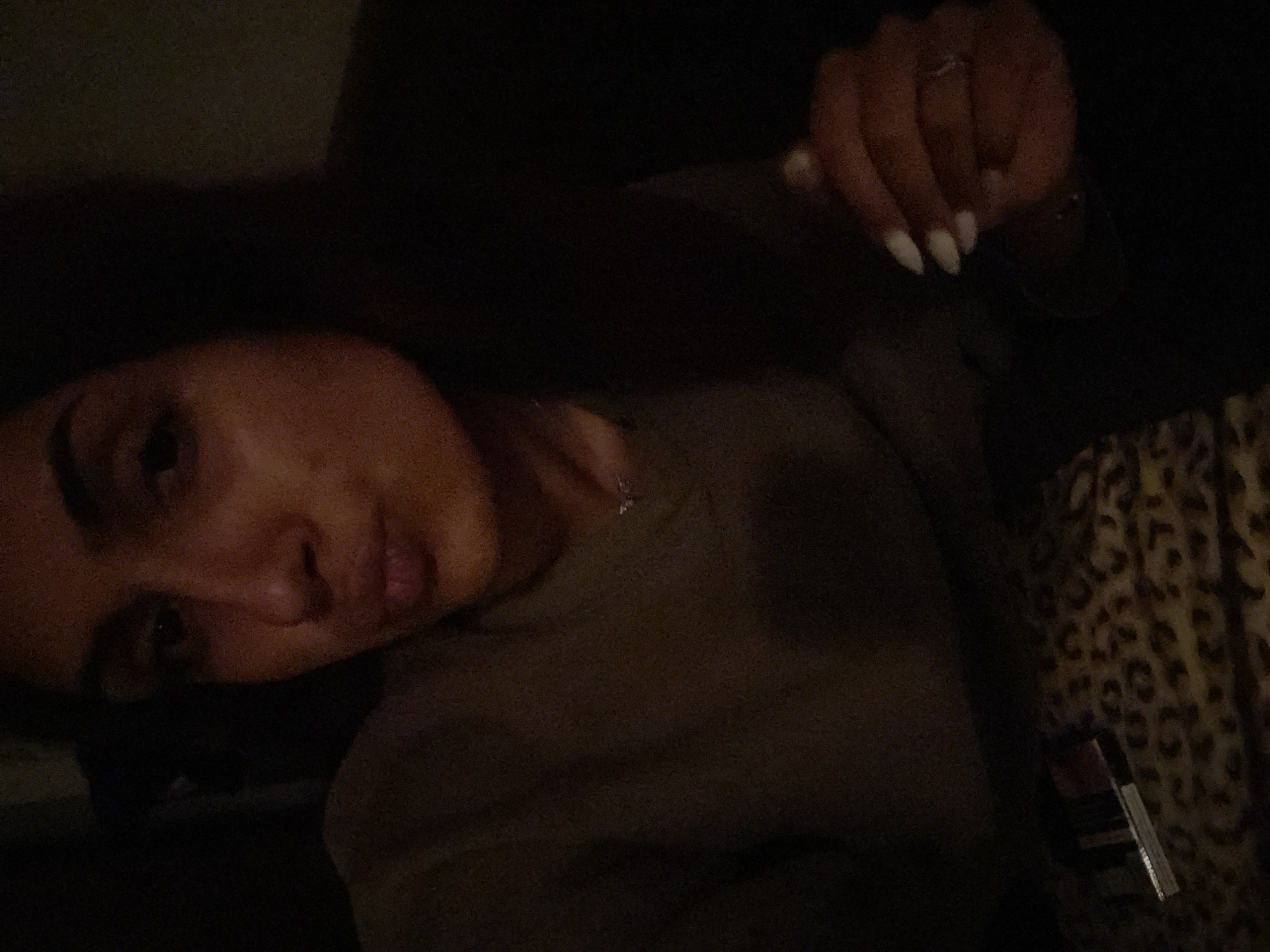 The image is a dimly lit, low-resolution selfie. The subject is a woman with dark hair, facing forward and puckering her lips. She is wearing a dark, long-sleeved shirt. A small, silver necklace is visible around her neck. Her right hand, with painted fingernails, is positioned in the lower right quadrant of the frame, with a ring on one of her fingers. In the lower left quadrant, a leopard-print surface is visible, with what appears to be a cigarette pack and some other small objects on it. The background is dark and indistinct. There is no discernible information within the image that would indicate a specific location in Varna, Bulgaria.