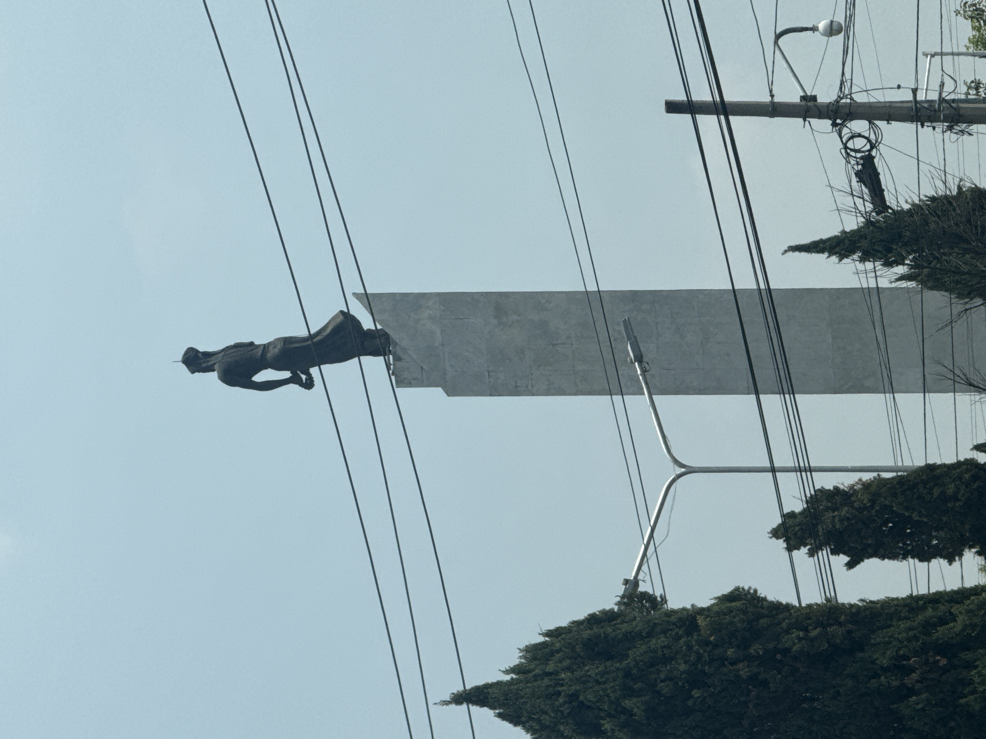 An eye-level, rotated image shows a statue atop a structure, against a blue sky. In the upper right and lower right corners are dark green leafy trees, possibly evergreens. Electrical power lines crisscross the image in straight, diagonal lines. In the upper right is a wooden power pole with a street lamp attached, along with a tangle of electrical equipment and wires. A statue of a man appears to be diving off the end of a gray, elevated platform. The monument appears to be located in Puebla, Mexico.