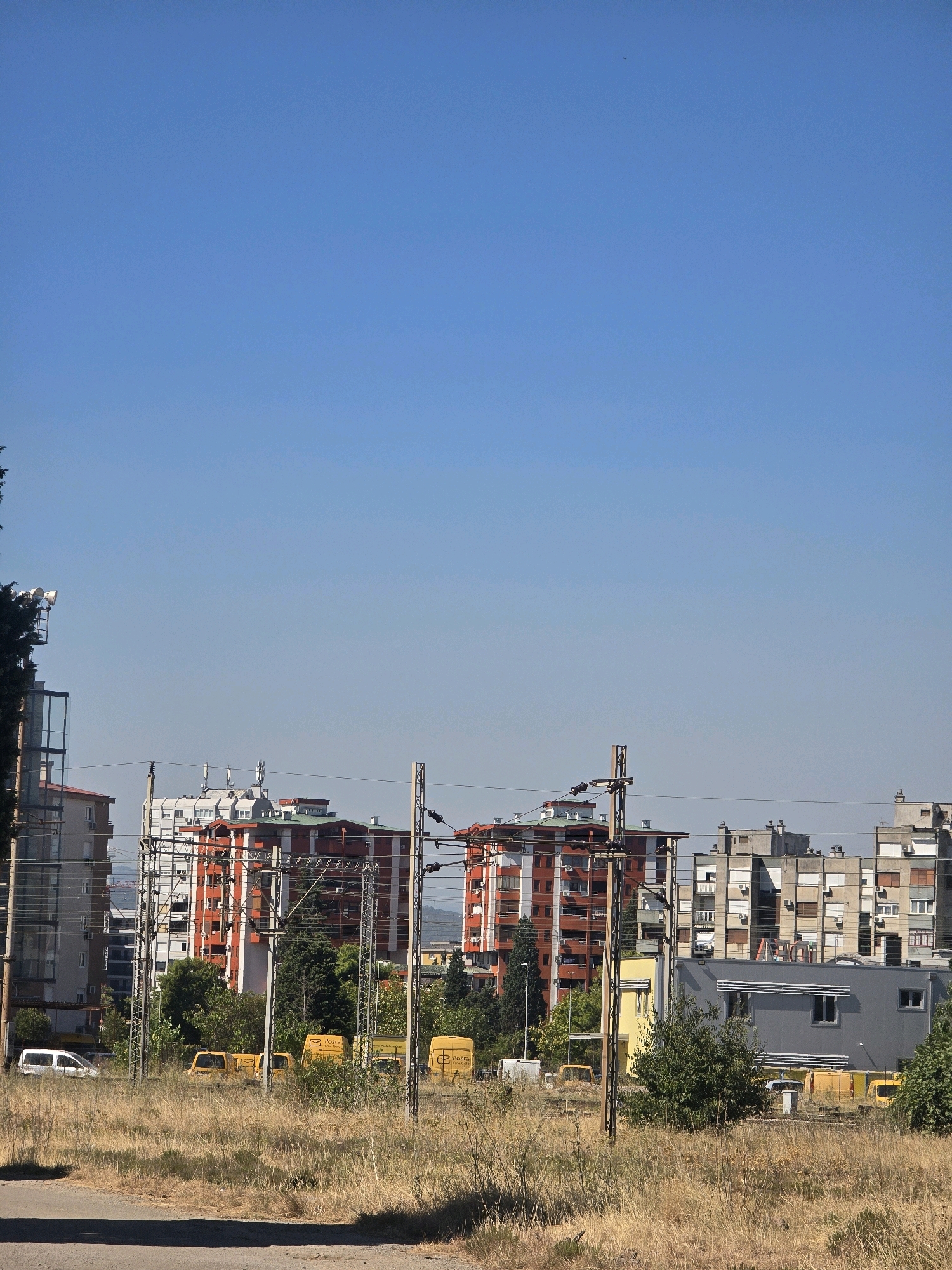The image shows a view of urban development in Podgorica, Montenegro. In the foreground, dry, tall grass covers the ground, with a gray asphalt path visible at the very bottom. Behind the grass, several yellow vans are parked, some with "Posta" branding, suggesting a postal service depot. A white van is also parked among them. In the middle ground, several residential and commercial buildings are visible. These buildings vary in height and architectural style, with some featuring orange and white facades, while others are more modern gray structures. Electrical poles and wires crisscross the scene, indicating an active power grid. The background is dominated by a clear blue sky. The overall impression is a functional, somewhat utilitarian urban landscape under a bright, sunny sky.
