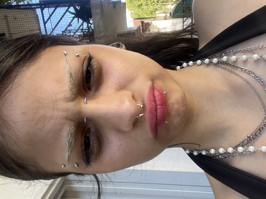 A woman is shown from a low angle, close-up. She has several facial piercings, including the eyebrows, nose, and lips, all of which have metal studs. She wears multiple silver necklaces and a black tank top. Her eyebrows appear to be bleached or painted white. Her expression appears stern, with a furrowed brow. The background features a blue metal structure, a building, and hints of greenery. The composition emphasizes her unique style and facial features.