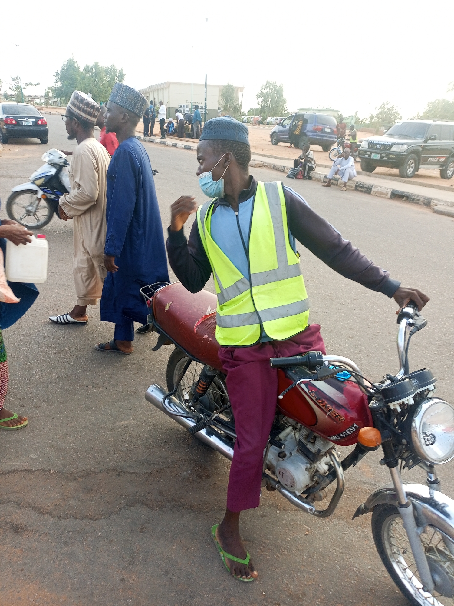 A man wearing a blue cap, a light blue shirt, a high-visibility yellow vest, and maroon pants is leaning against a red motorcycle on an asphalt road. He is also wearing green flip-flops. The motorcycle has a silver headlight and a chrome exhaust pipe. He is wearing a blue surgical mask.
Behind him, two men walk in the same direction, one in a light brown robe with a patterned cap, and the other in a blue robe and patterned cap. A white motorcycle is visible between them. A woman holding a white container is partially visible to the left.
In the background, there are several parked vehicles including a black car, a blue van, and a black SUV. Several people are sitting or standing near the parked vehicles. A light-colored building can be seen in the distance.
The scene appears to be outdoors on a sunny day. The image is taken at ground level, on a paved road in the city of Gombe, Nigeria.
