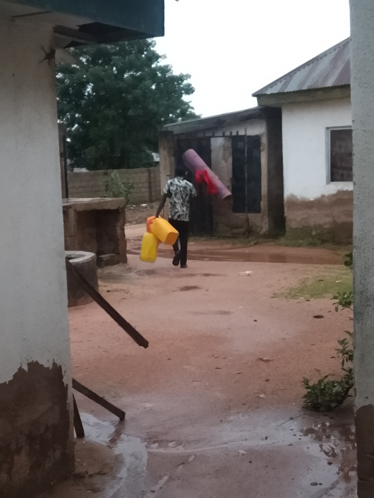 In an outdoor, dusty environment, a person is walking away from the camera, carrying two yellow containers in their left hand and a rolled-up mat or blanket in their right hand. The person is wearing a dark and white patterned shirt and dark pants. Behind the person, there is a building with a corrugated metal roof, and a large tree with dark green foliage. The ground appears to be a mixture of dirt and scattered puddles, suggesting recent rain or a water source. To the left of the frame, part of a white building with some discoloration is visible. The overall scene suggests a somewhat rural or undeveloped area.