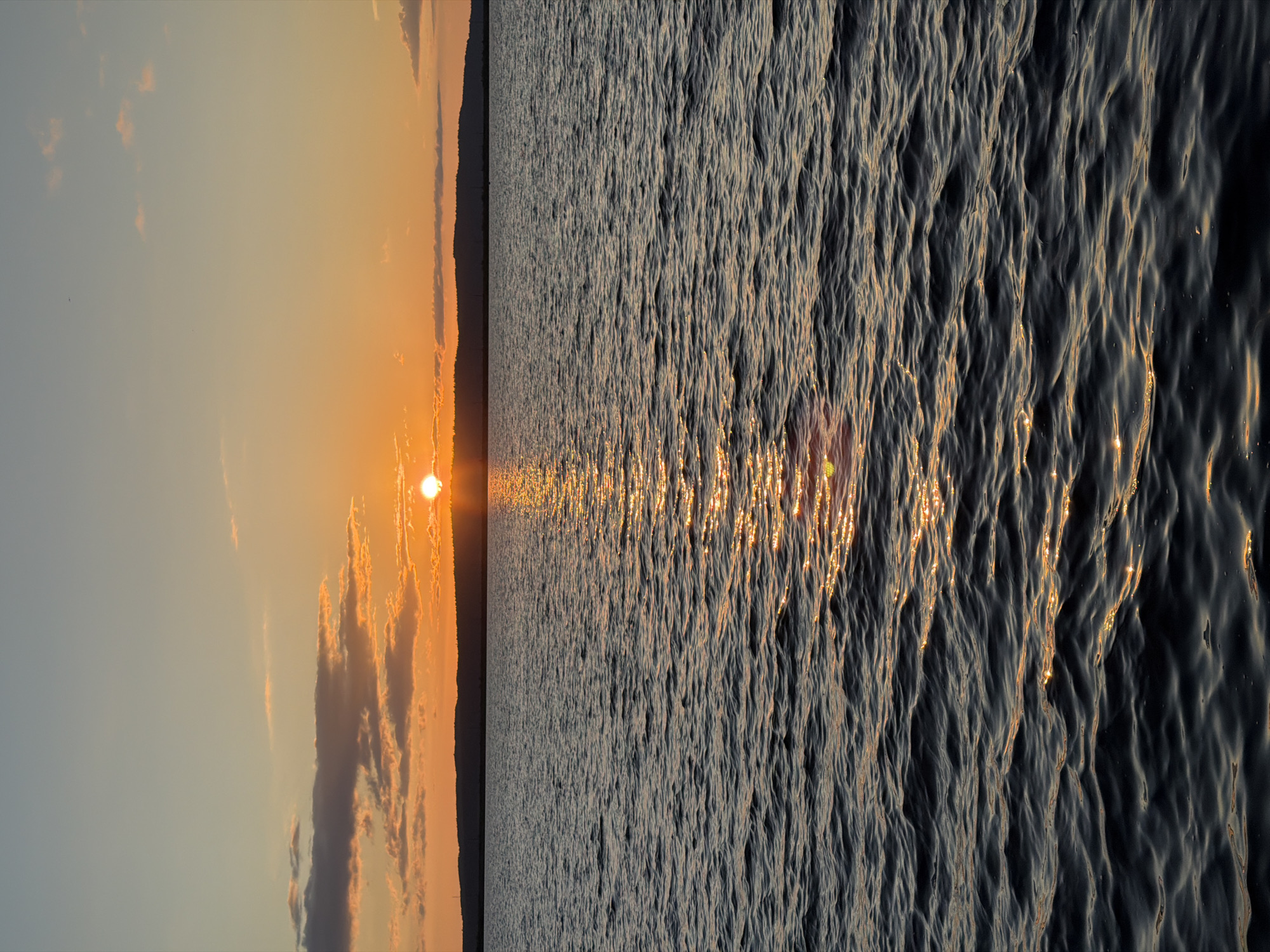 The image depicts a sunset over a large body of water, likely the Kama River in Chaikovsky, Russia. The sun, a brilliant orange orb, is positioned just above a dark, silhouetted landmass on the horizon. The sky transitions from a soft, pale blue at the top to warm hues of orange and yellow near the sun, with scattered, wispy clouds catching the light. A shimmering reflection of the sun streaks across the rippling surface of the water, creating a pathway of golden light. The water itself is dark and textured with small waves. The scene is devoid of people, structures, or any other discernible objects, focusing solely on the natural landscape.