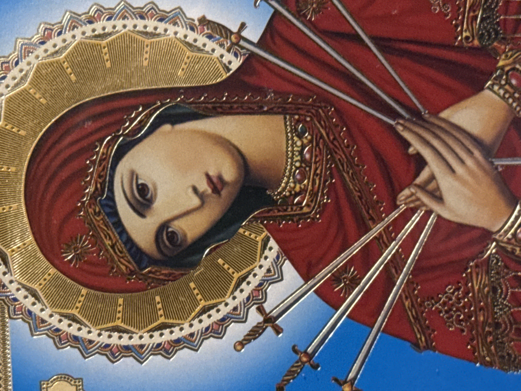 The image is a close-up, vertically oriented, full-color, studio photograph of an icon of the Virgin Mary, or Madonna. The icon features the Virgin Mary's face and upper torso. She is wearing a red veil and a red garment. Seven swords are depicted piercing her chest from left to right, with their hilts visible. The swords are metallic and appear to be antique or ceremonial. The Virgin Mary's hands are clasped together in front of her chest, in a gesture of prayer or sorrow. She has a halo behind her head, with a detailed gold and white circular design. The background is a solid, bright blue. The icon is ornately decorated with gold trim and colorful embellishments. The image is sharp and well-lit, capturing the details of the artwork.
