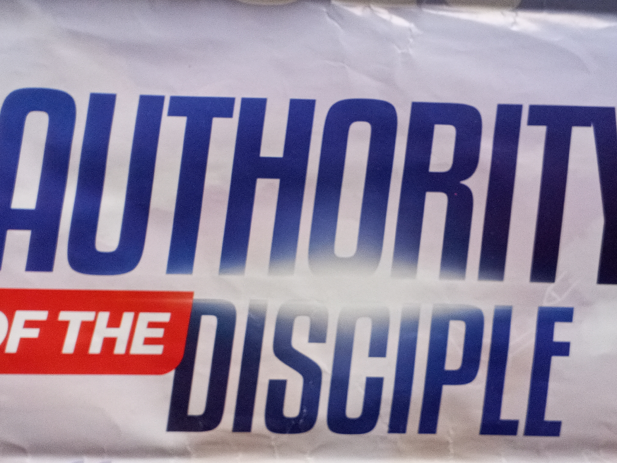 The image shows a close-up of a banner with the words "AUTHORITY OF THE DISCIPLE." The word "AUTHORITY" is in large, blue, sans-serif font. Below it, "OF THE" is in white lettering on a red rectangular background. To the right of that, "DISCIPLE" is in a similar blue, sans-serif font as "AUTHORITY," but slightly smaller. The banner appears slightly wrinkled. The location of the city of Badagry, Nigeria, is not discernible from the image.