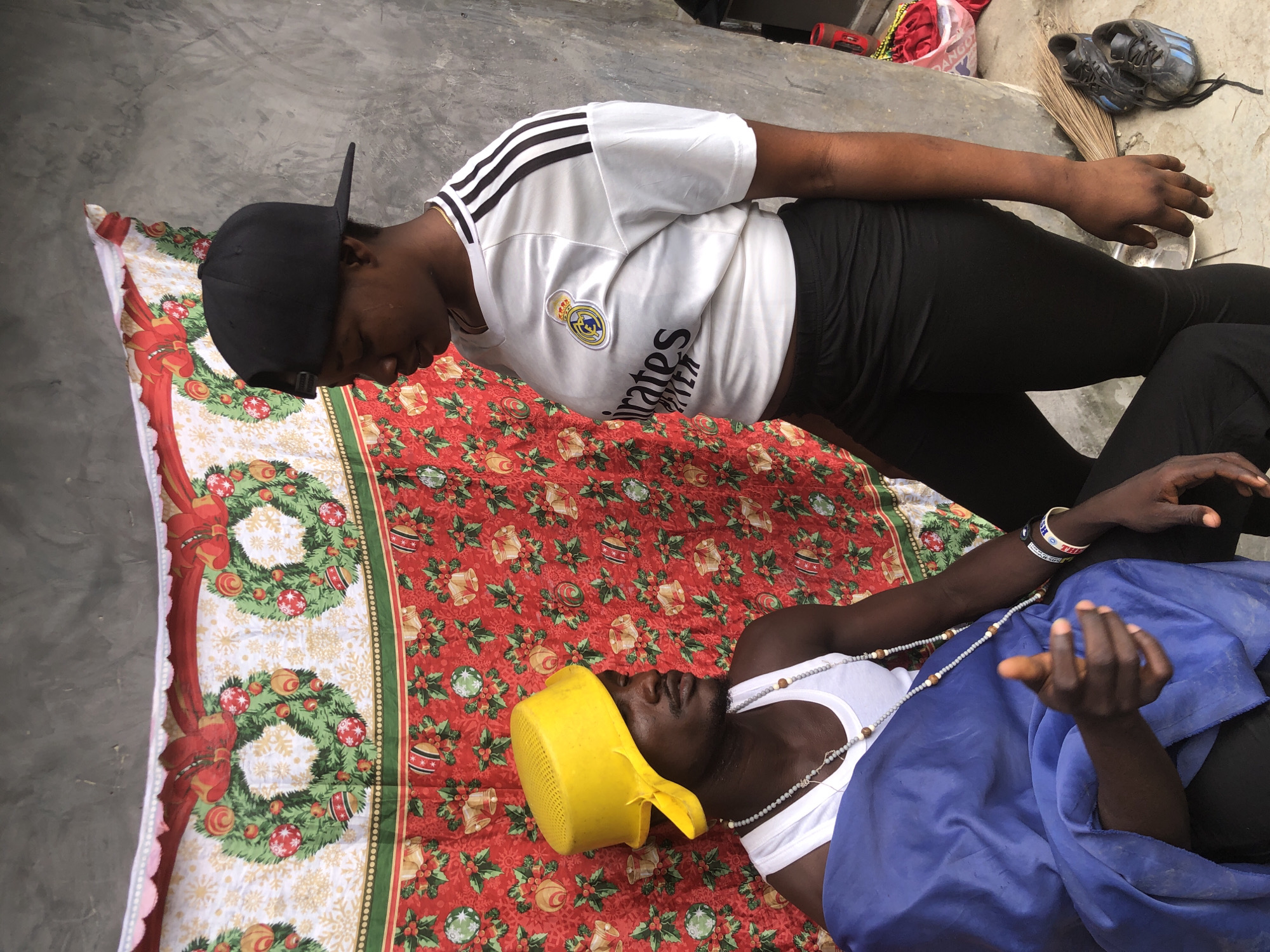 In the city of Ado, Nigeria, a man and a woman are positioned in front of a Christmas-themed backdrop. The man, wearing a white tank top and a blue cloth draped over his lap, has a yellow colander on his head and is adorned with necklaces. He is looking to his left with his eyes closed. The woman, on the right, is wearing a white Real Madrid jersey with black stripes on the shoulders and a black baseball cap. She is looking down at the man. The backdrop features festive Christmas wreaths, red holly berries, and green leaves. To the right of the subjects, a broom and a pair of black and blue sneakers are visible on the ground. The overall scene appears to be an outdoor setting with a concrete wall in the background.
