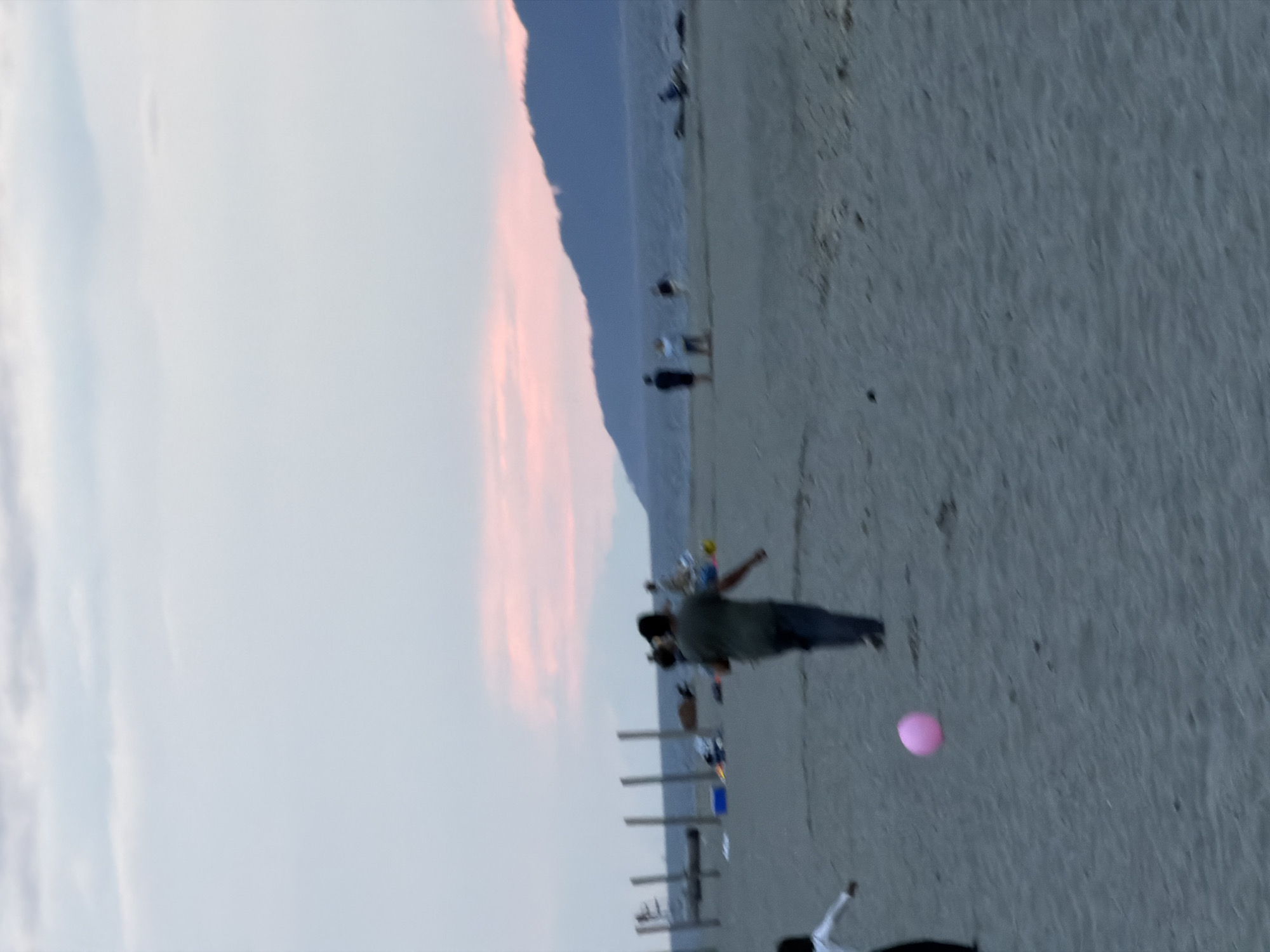 The image captures a beach scene at dusk, likely in Vancouver, Canada, given the mountainous backdrop typical of the region. In the foreground, a person in a dark shirt and pants is walking on the sand, seemingly kicking or interacting with a pink balloon. To the left, another figure, partially visible and dressed in white, is also on the beach. In the mid-ground, several other people are dispersed across the sand, some standing and others appearing to be sitting near beach structures, possibly driftwood or pilings. The water stretches out to the horizon, where a dark mountain range is silhouetted against a soft pink and pale blue sky, indicating the setting sun. The overall impression is of a casual, late-day gathering on a sandy beach.