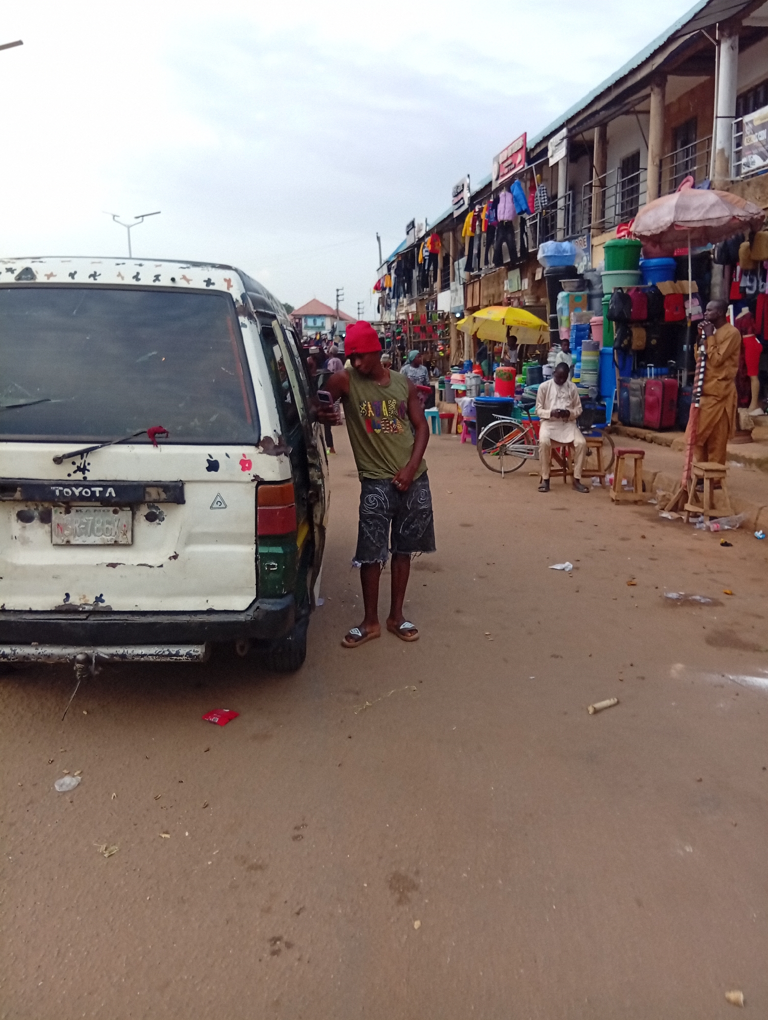 In Bukuru, Nigeria, a street market scene unfolds under an overcast sky. A white Toyota minivan, heavily weathered with rust and peeling paint, is parked on the left, its rear end angled towards the viewer. Its license plate, partially obscured, reads "NSK-786X". To the right of the van, a young man wearing a red cap, a green tank top with a colorful graphic, and denim shorts stands, looking at his phone. He wears sandals.
The background reveals a row of market stalls lining a street. Various goods are displayed, including stacked plastic containers in bright colors, clothing hanging on display, and umbrellas, one a cheerful yellow, another a larger, faded pink. A man in a light-colored tunic is seated on a stool, looking down at his phone, with a bicycle parked beside him. Further down the street, another man in a brown garment stands. The ground is a dirt and asphalt surface, littered with debris, including a red packet and scattered organic matter. The overall impression is a busy, functional marketplace.