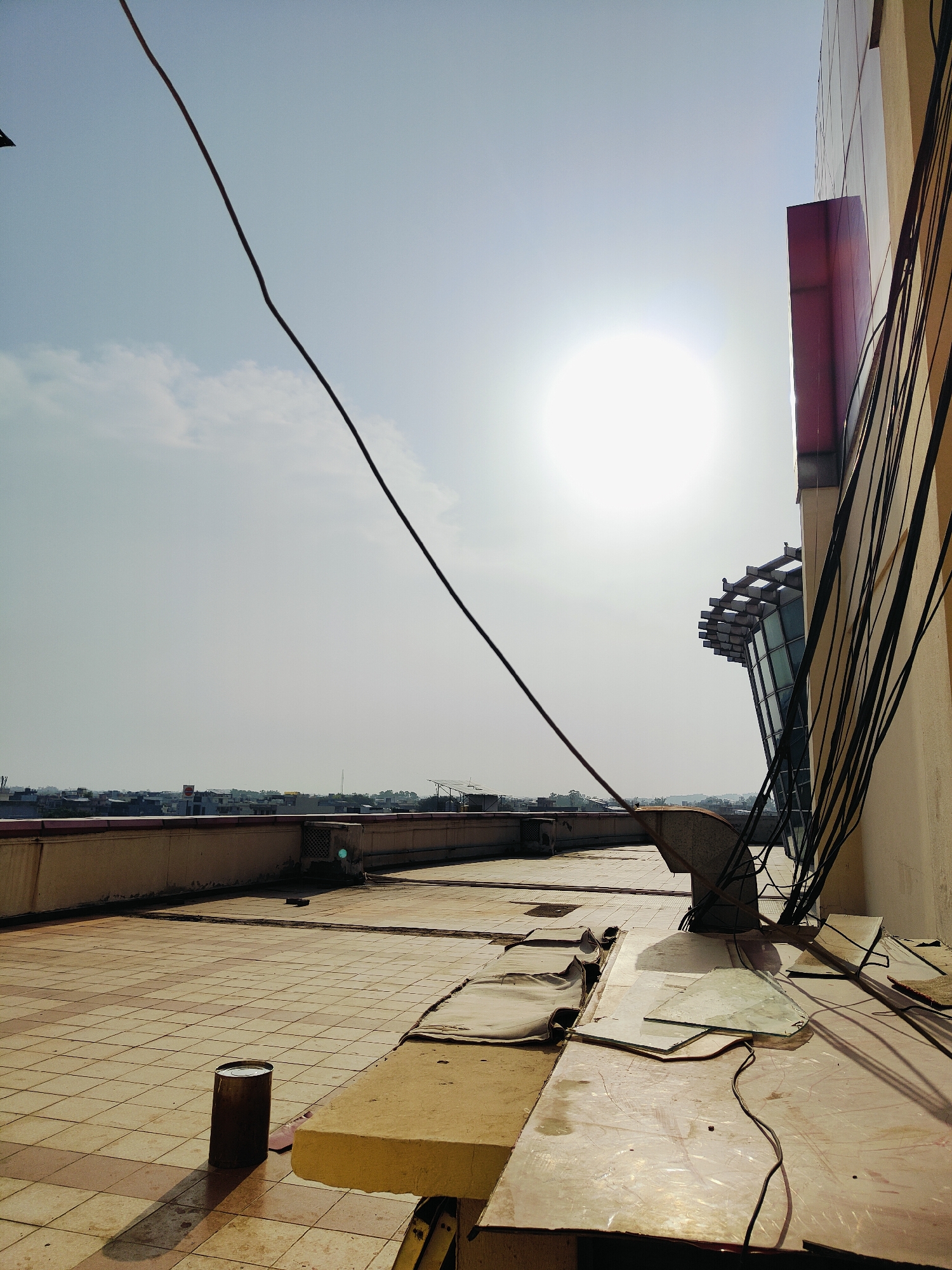 The image depicts a rooftop scene in Gurgaon, India, on a bright, sunny day. The foreground is dominated by a tiled floor, with a large, metallic cylindrical object, possibly a drum or bin, casting a long shadow. To the right, a section of a building's exterior wall, painted a light beige, is visible, with a prominent red rectangular advertisement or design element. A bundle of black electrical wires runs vertically along the side of this building.
Further into the scene, a modern architectural structure with glass windows and a cantilevered roof design juts out from the building. On the rooftop, various construction or maintenance materials are scattered, including what appear to be sheets of glass, rolled-up tarpaulin or insulation, and a large grey ventilation duct. In the background, a low-rise urban skyline of Gurgaon can be seen under a hazy sky, with hints of residential or commercial buildings and some vegetation. The sun is a bright, almost blinding orb in the upper center of the sky. The overall impression is one of an urban rooftop undergoing work or maintenance, with the characteristic environment of an Indian city visible in the distance.