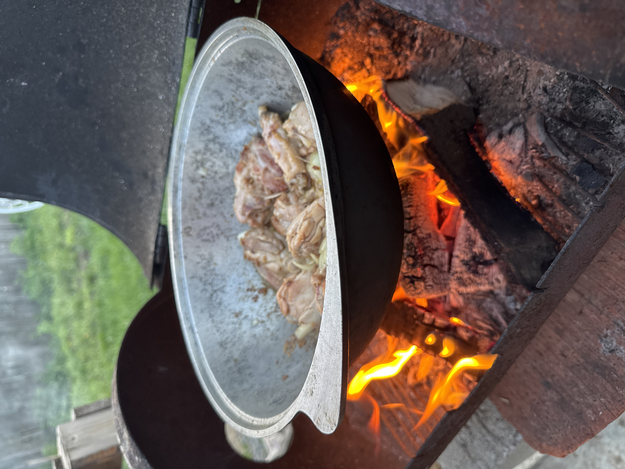 A metal cauldron containing raw, seasoned chicken pieces is positioned over a wood-fueled fire. The fire burns brightly with visible flames and partially charred wood. The cauldron is placed in a stove/fire pit. The setting appears to be outdoors, as suggested by the presence of greenery in the background on the left side of the picture. The image was taken in a location inside of a forest, probably in the city of Chaikovsky, Russia. The picture has been taken by an unknown person from a high angle.