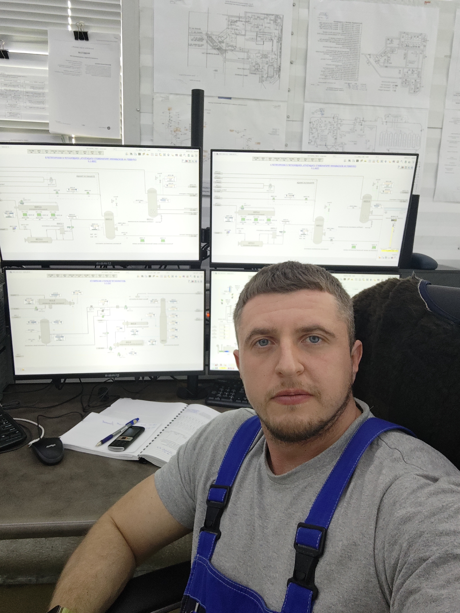 A man is shown in an indoor environment, likely an industrial control room, as he is sitting at a desk surrounded by four computer monitors displaying graphical representations of industrial processes. The walls behind the monitors are covered with engineering schematics and documents. On the desk, there is a mouse, a notebook, a pen, and an old cell phone. The man is wearing a gray t-shirt with blue overalls, and he is looking directly at the camera. The location is unknown.