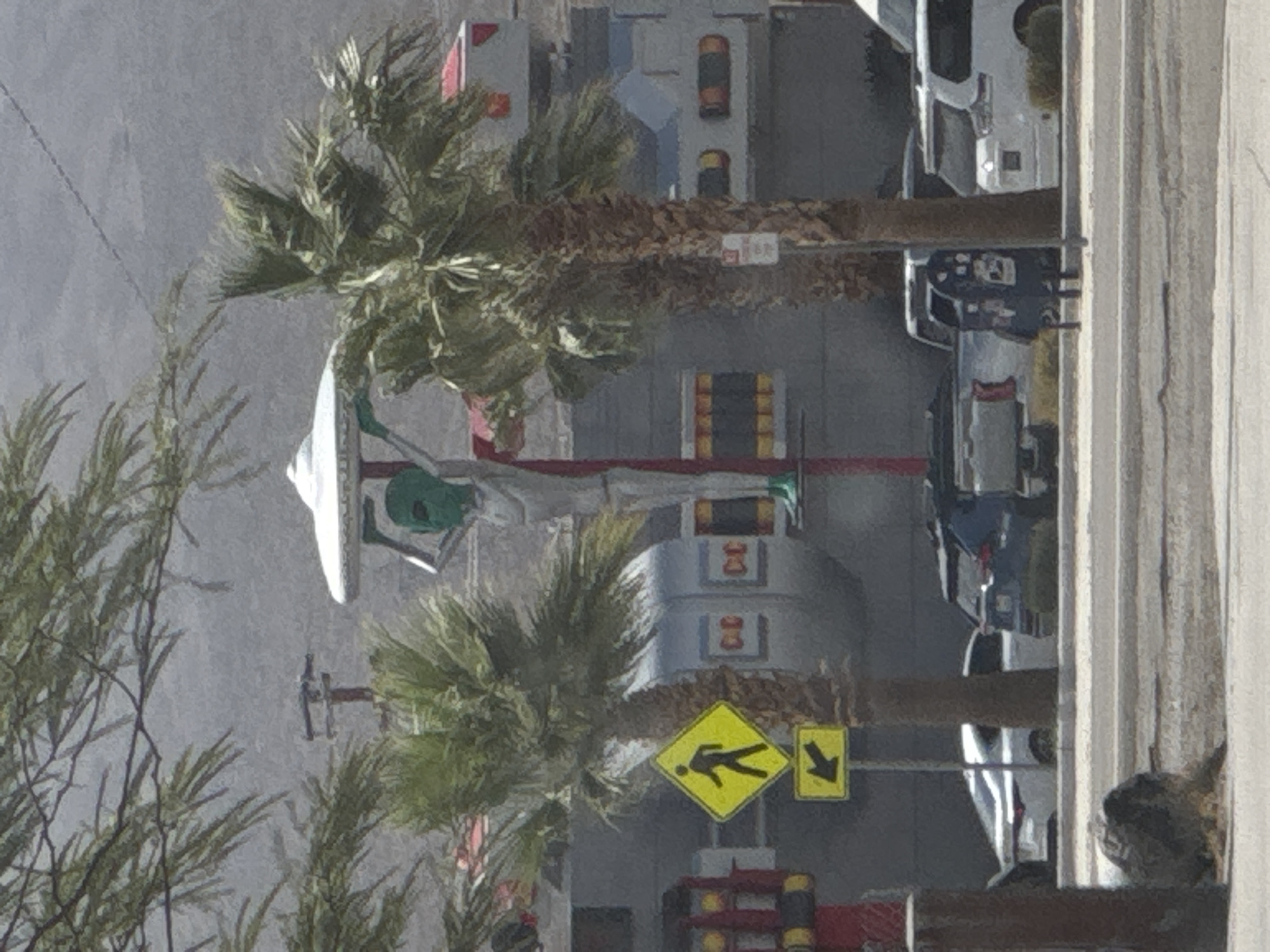 The image depicts a scene in Baker, California, United States of America. The central focus is a large, silver, alien-shaped statue holding a white, saucer-like object above its head. The statue is positioned in front of a building complex with a grey facade and red accents. Two palm trees are visible in the foreground, one on either side of the alien statue. To the left of the alien, a yellow diamond-shaped pedestrian crossing sign with a black silhouette of a person walking is mounted on a pole, with a smaller yellow rectangular sign indicating a left turn below it. In the background, several vehicles are parked, including a white sedan, a dark blue SUV, and a white van. The environment appears arid, with dry vegetation and a mountainous landscape visible in the distance.