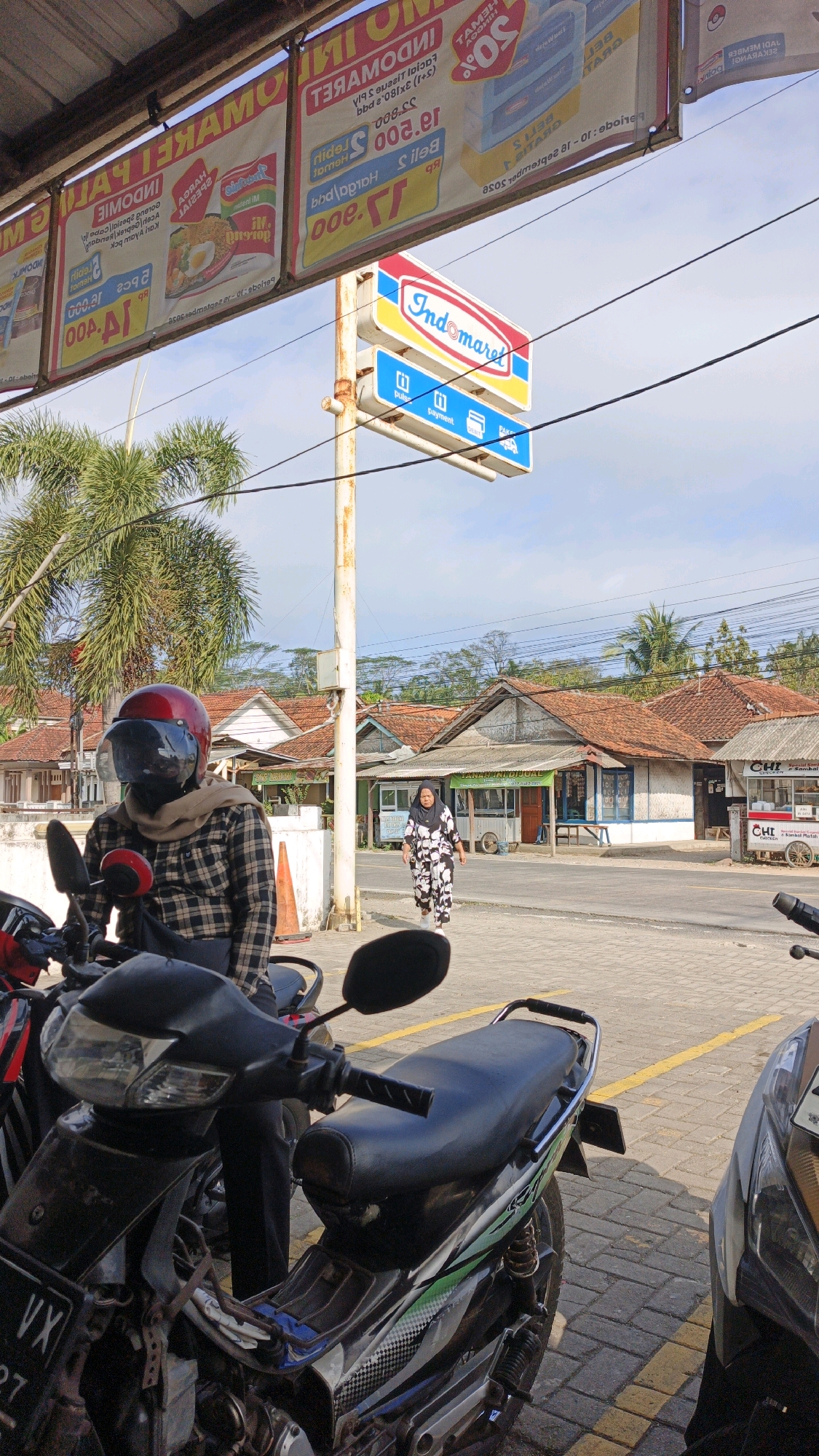 The image depicts an outdoor scene in Cibenda, Indonesia. In the foreground, several motorcycles are parked. One motorcycle in the center is partially visible, showing its black seat, handlebars, and parts of its engine. A person wearing a red helmet and a plaid shirt is seated on a motorcycle to the left, partially obscuring it. Behind the parked motorcycles, a street runs parallel to a row of small shops with tiled roofs. An Indomaret convenience store sign is prominently displayed on a pole. A woman wearing a black and white patterned dress is walking on the sidewalk in front of the shops. Banners advertising promotions are hanging overhead. The sky is partly cloudy.
