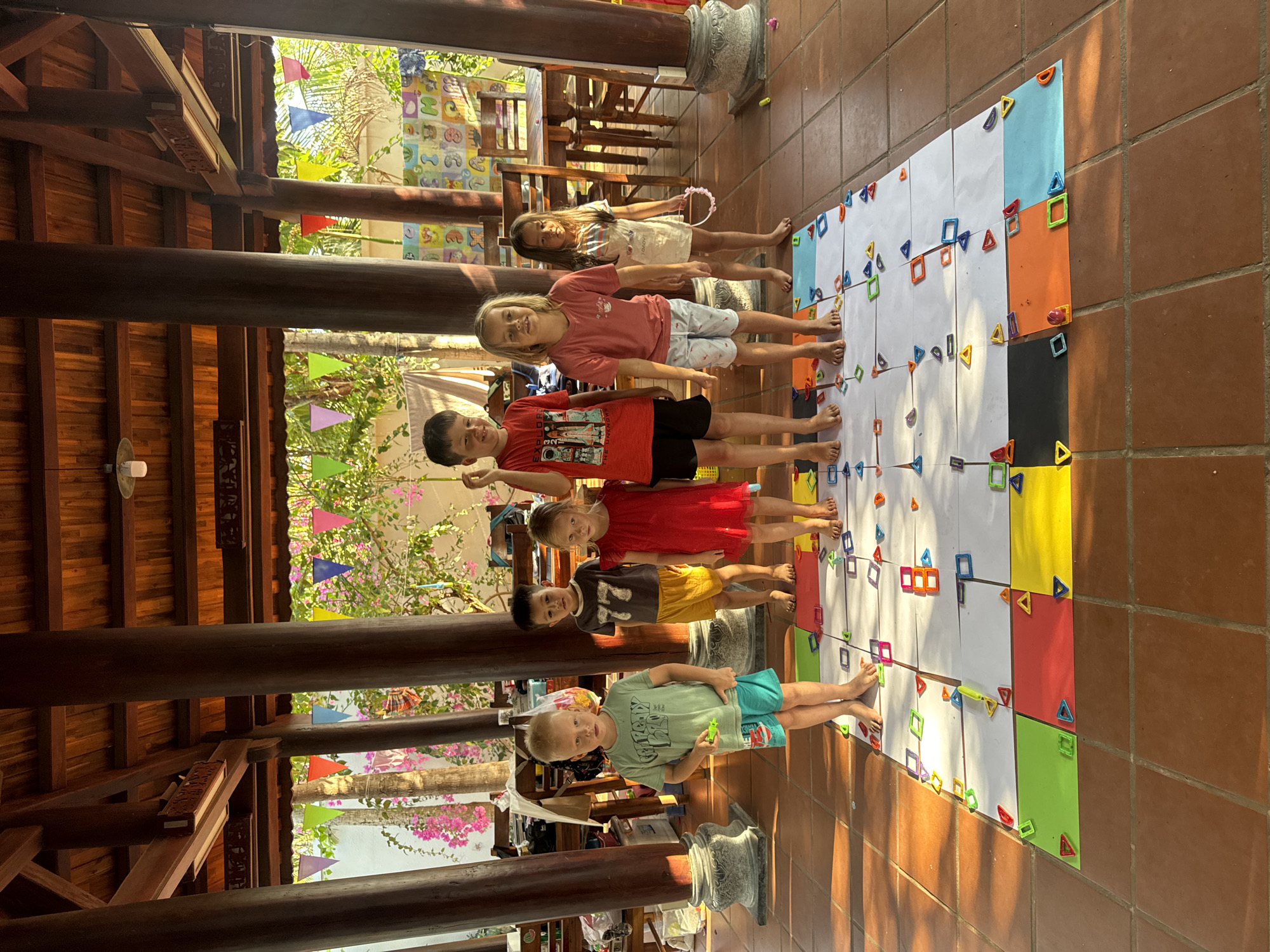 Seven children are captured standing barefoot in a line on a large, colorful game mat laid out on a terracotta-tiled floor. The mat is composed of white and brightly colored paper squares (yellow, red, green, blue, orange, black), adorned with numerous magnetic tiles (squares and triangles) in various hues, arranged into patterns or paths.
The children, of diverse ages and appearances, face the camera, many with smiles or relaxed expressions. From left to right: a blonde boy in a light green t-shirt and blue shorts holds a small green toy; next is a dark-haired boy in a grey '77' t-shirt and yellow shorts; followed by a blonde girl in a red dress; then a dark-haired boy in a red t-shirt that prominently displays "EXPLORE THE UNKNOWN" and black shorts; a blonde girl in a pink t-shirt and patterned shorts; and a dark-haired girl in a light dress holding a pink hoop. The legs of another child are partially visible further along the line.
The setting is an open-air or semi-covered area, possibly a pavilion or veranda, featuring robust dark wooden beams and columns, some with intricate carvings, suggesting traditional Vietnamese architecture. Lush green foliage, trees, and vibrant pink bougainvillea are visible beyond the structure under bright, natural sunlight, which casts distinct shadows on the tiled floor, indicating it is daytime, likely a clear, sunny late morning or early afternoon. Colorful triangular flags are strung among the background greenery. The scene portrays a cheerful and relaxed atmosphere, with the children seemingly engaged in an interactive game or learning activity. This environment is consistent with the specified location of Mũi Né, Vietnam.