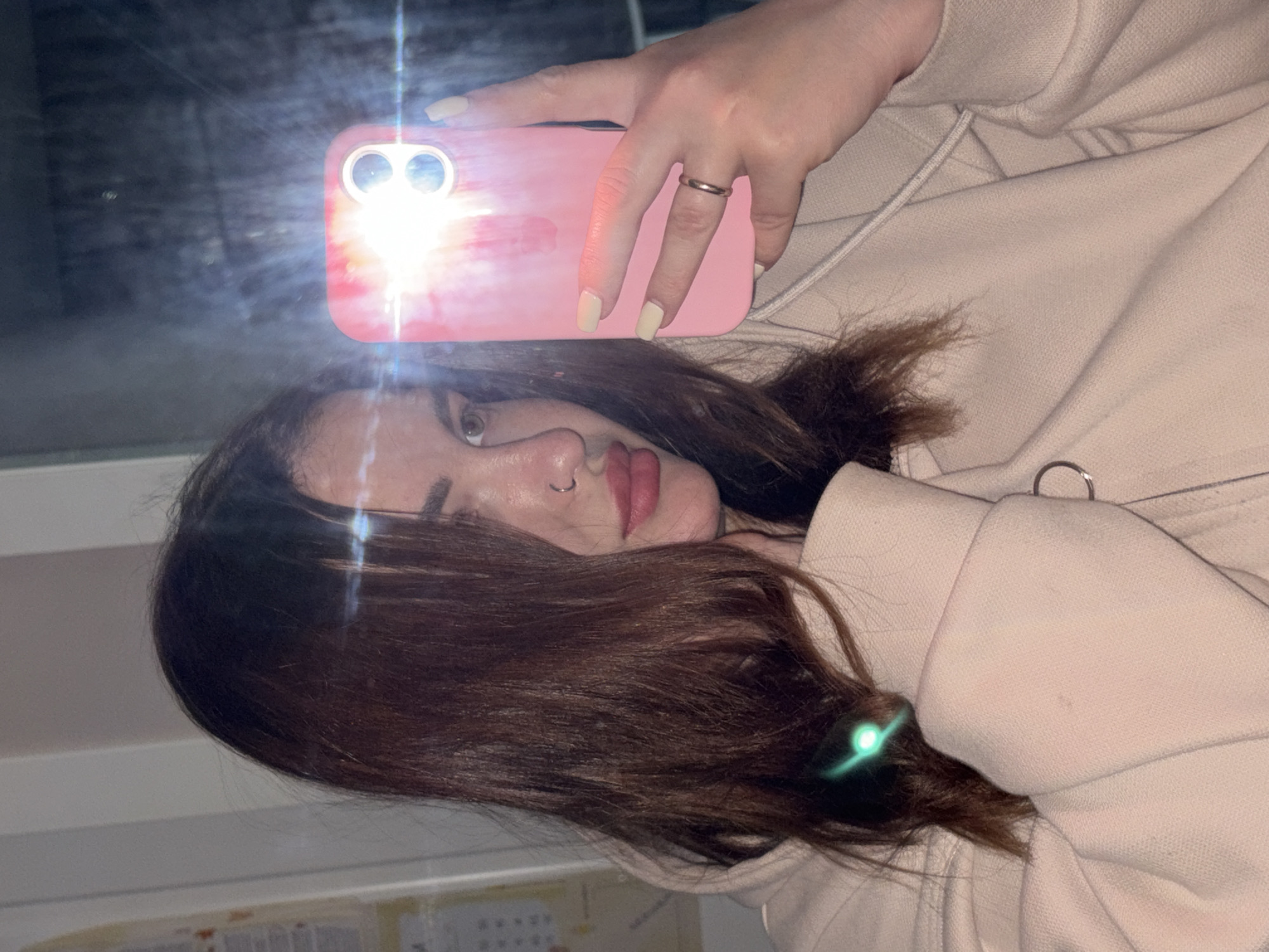 A woman with brown hair takes a selfie using a pink phone with the flash on. She has a ring in her nose and is wearing a ring on her finger. She's wearing a light-colored hooded sweatshirt. The background includes a window. I cannot confirm the location.