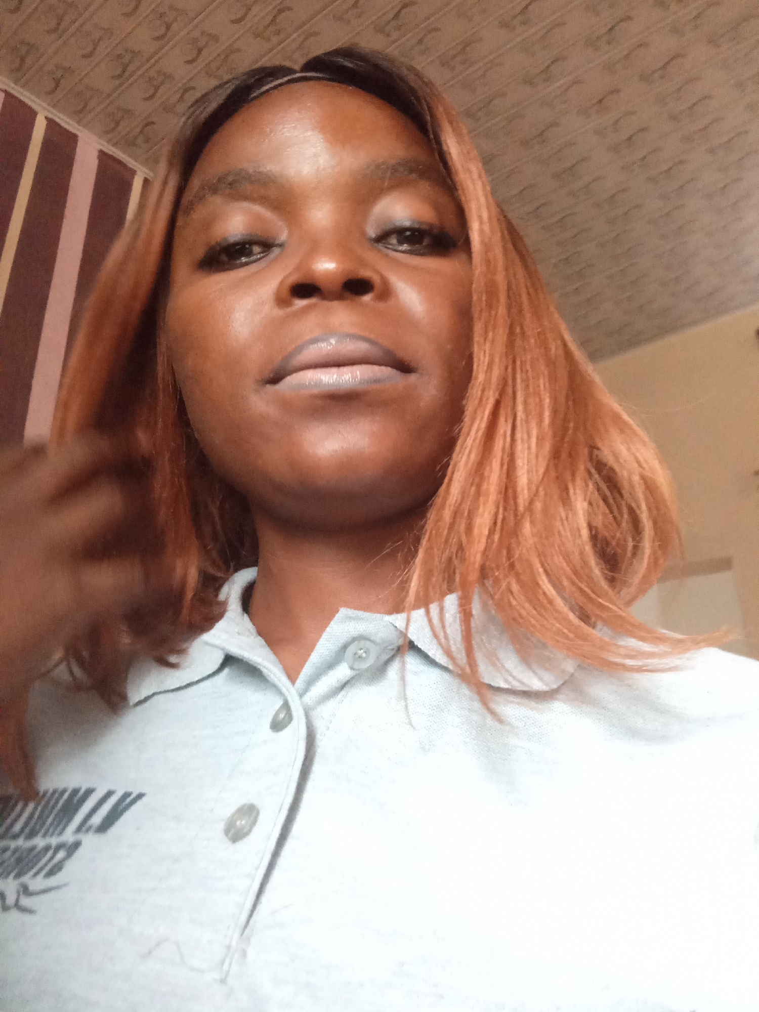 The image is a close-up, low-angle selfie of a Black woman with medium-toned skin and reddish-brown hair. She is wearing a light blue collared shirt with buttons. The background appears to be an indoor setting with a textured ceiling and a wall with striped wallpaper. There is text visible on the woman's shirt, which is partially obscured. The overall lighting suggests it is daytime. There is no information in the image to indicate the location is the city of Jalingo, Nigeria.