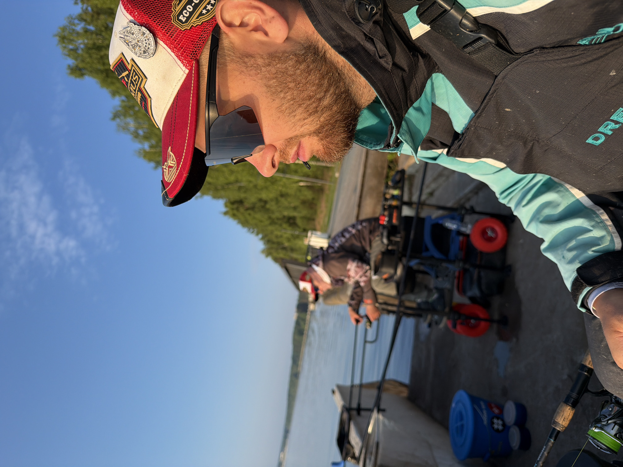 An adult male wearing sunglasses, a baseball cap, and a fishing vest is shown in a close-up shot, looking out at a body of water. He has a beard and is standing outdoors. In the background, another person is visible, also engaged in fishing. Fishing rods and equipment are present. The environment appears to be a waterfront with trees and a clear blue sky. The scene is set during daylight hours. There is no indication of the specific location within Chaikovsky, Russia.