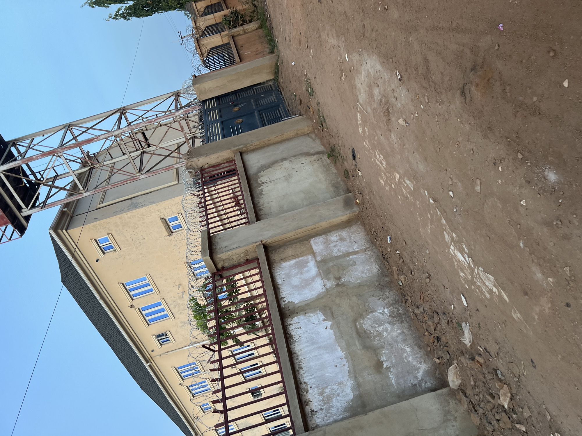 The image depicts a multistory building in Abuja, Nigeria, with a beige facade and blue-tinted windows. A metal scaffolding structure extends from the building's side. The building is surrounded by a concrete structure that has some paint. A barbed wire fence is visible. The adjacent ground is an unpaved dirt path. The perspective is from a low angle, and the image is slightly rotated. No people are visible.