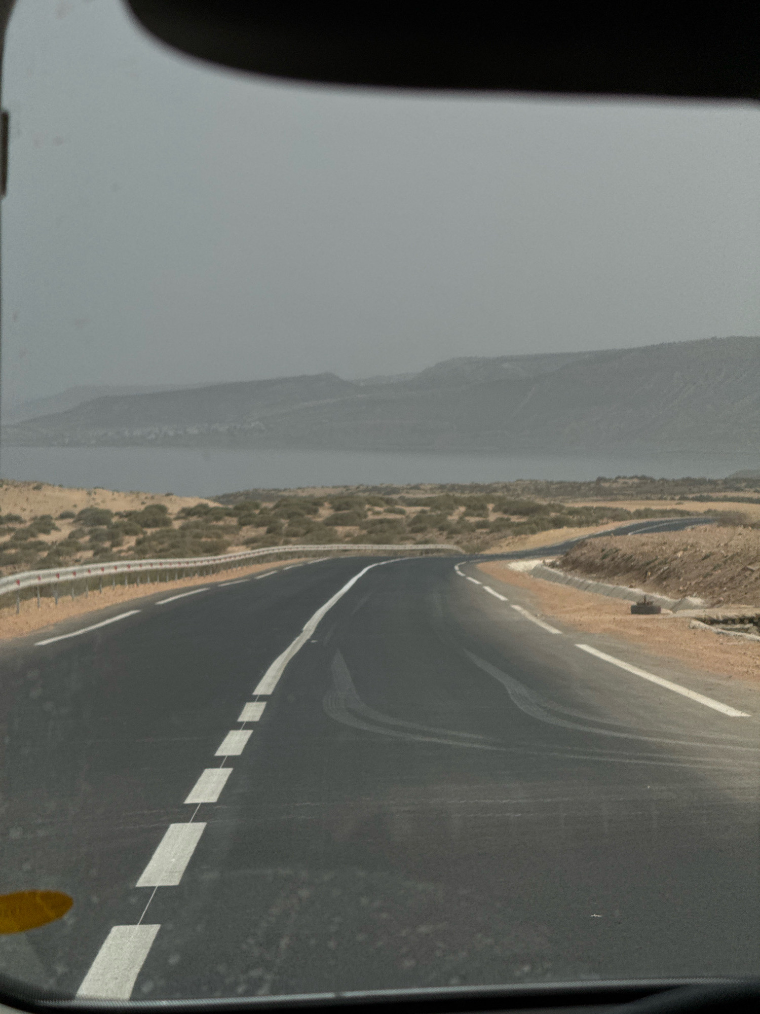The image, taken from inside a vehicle, shows a two-lane asphalt road curving to the right. White dashed lines mark the center of the road, and a solid white line is on the right edge of the road. The road is bordered by a sandy shoulder with sparse, low-lying green vegetation and a metal guardrail. In the background, a body of water, possibly an ocean or a large lagoon, is visible, separated from the road by more arid land with scrub brush. Beyond the water, hazy, undulating hills form the horizon under an overcast sky. The scene is consistent with the coastal landscape near Tamri, Morocco, known for its dry climate and sparse vegetation. There are no people or visible animals in the image. The overall impression is one of a quiet, somewhat desolate, but scenic route.
