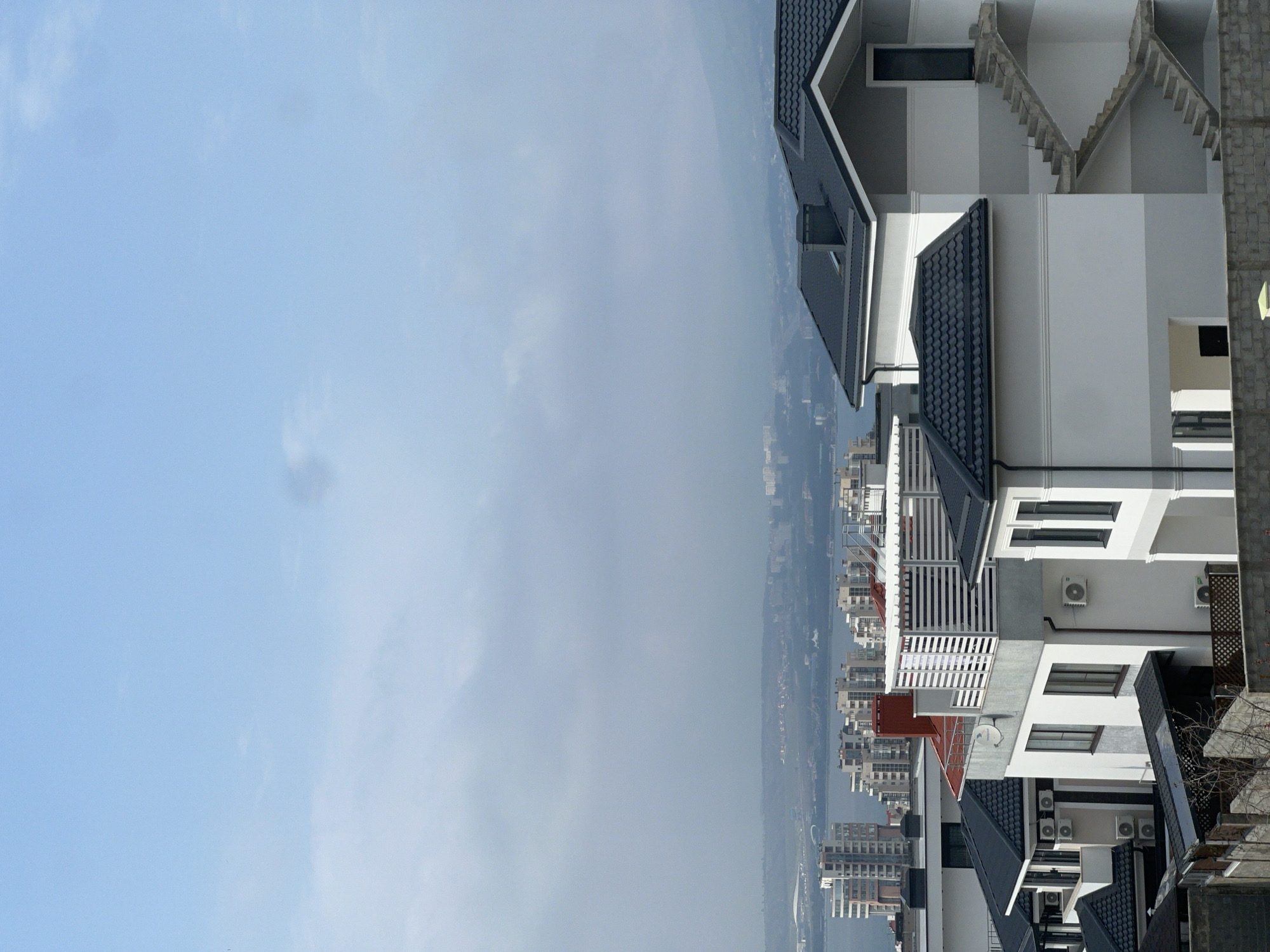 The image presents an elevated, panoramic view of a residential district in Gelendzhik, Russia. In the foreground and midground, several modern, multi-story houses are visible, characterized by dark gray pitched roofs and facades primarily composed of white and light gray sections. Exterior air conditioning units are affixed to the walls of multiple residences. A prominent feature on the far-right building is an external concrete staircase ascending to an upper-level entrance. One house displays a flat roof section equipped with a white slatted pergola or shade structure. A reddish-brown chimney protrudes from another rooftop. Satellite dishes are present on various building exteriors. The immediate foreground is defined by a low, light gray concrete block wall.
Beyond the cluster of houses, a significant body of water, identifiable as Gelendzhik Bay, stretches horizontally across the scene. The opposite shoreline is populated with numerous multi-story buildings, forming a distant urban landscape. Hazy mountains or hills rise behind these structures, blending into the distant horizon. The sky is overcast, exhibiting shades of light blue and gray with indistinct cloud formations. No individuals, explicit actions, or interactions are observed in the scene.