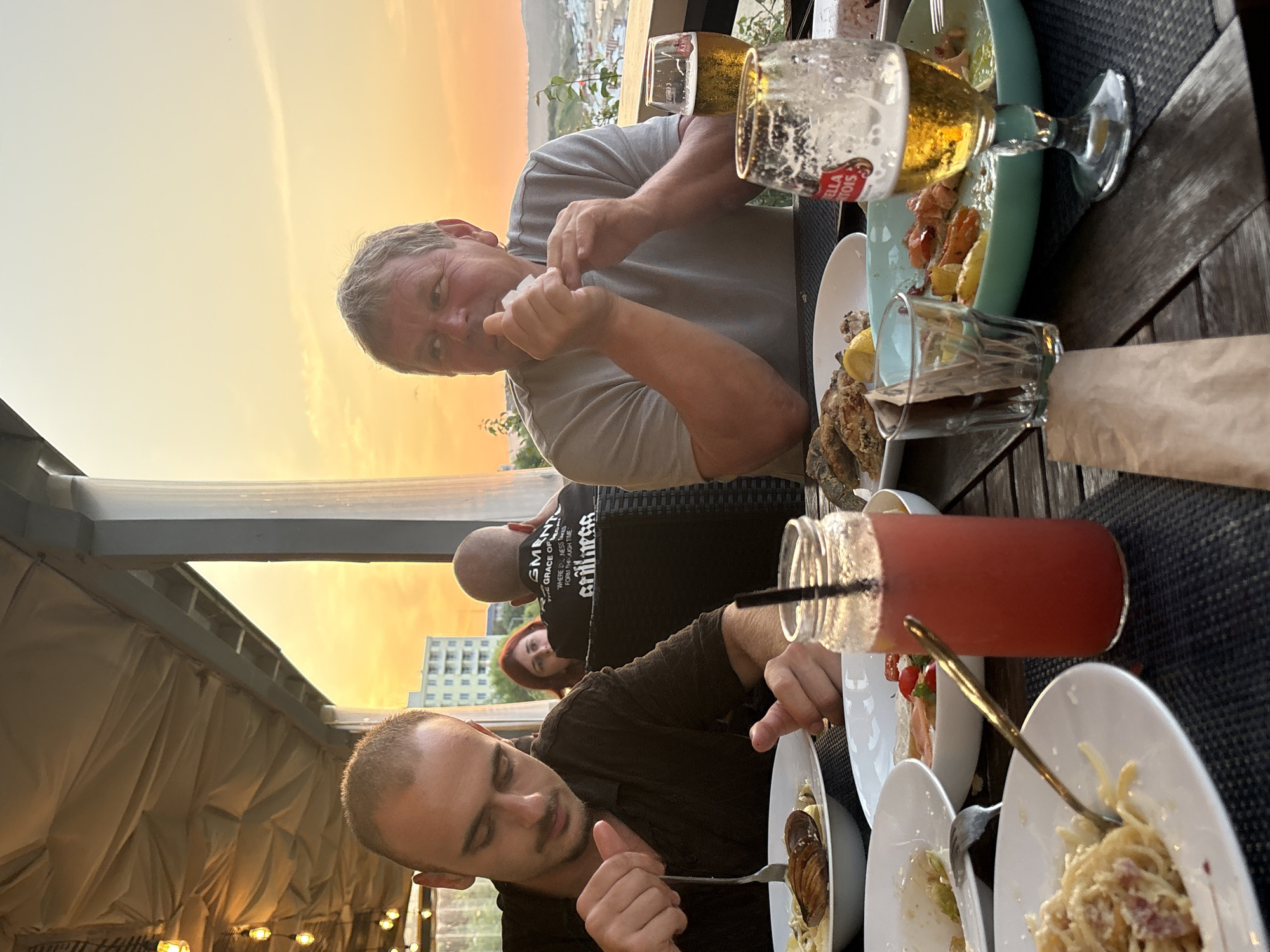 The image shows a group of people dining at an outdoor restaurant with a view of Primorsko, Bulgaria, during sunset. In the foreground, a man with short hair and a mustache is eating pasta with a fork. To his right, a glass jar filled with a red beverage and a straw sits on the table. Further to the right, a man with graying hair is wiping his mouth with a napkin while looking away from the camera. Two glasses of beer are on the table, one partially consumed, with a Stella Artois label visible. Plates of food, including pasta and what appears to be seafood, are scattered across the wooden table. In the background, a woman with red hair is visible, and further back, a man wearing a black t-shirt with text on the back is seated. The restaurant appears to be covered by an awning, with string lights visible overhead. The setting sun casts a warm, orange glow over the scene, illuminating buildings and hills in the distance, indicative of the coastal town of Primorsko.