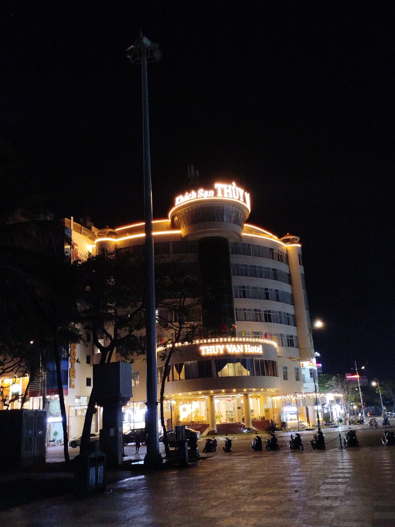 The image depicts the illuminated facade of the Thuy Van Hotel at night in Vung Tau, a coastal city in Vietnam near Ho Chi Minh City. The multi-story building features lighted signage, horizontal bands of light, and a prominent circular structure on top. A tall utility pole stands in the foreground. The street in front of the hotel has parked motorbikes and pedestrians. Other buildings and streetlights are visible in the background. The sky is dark.