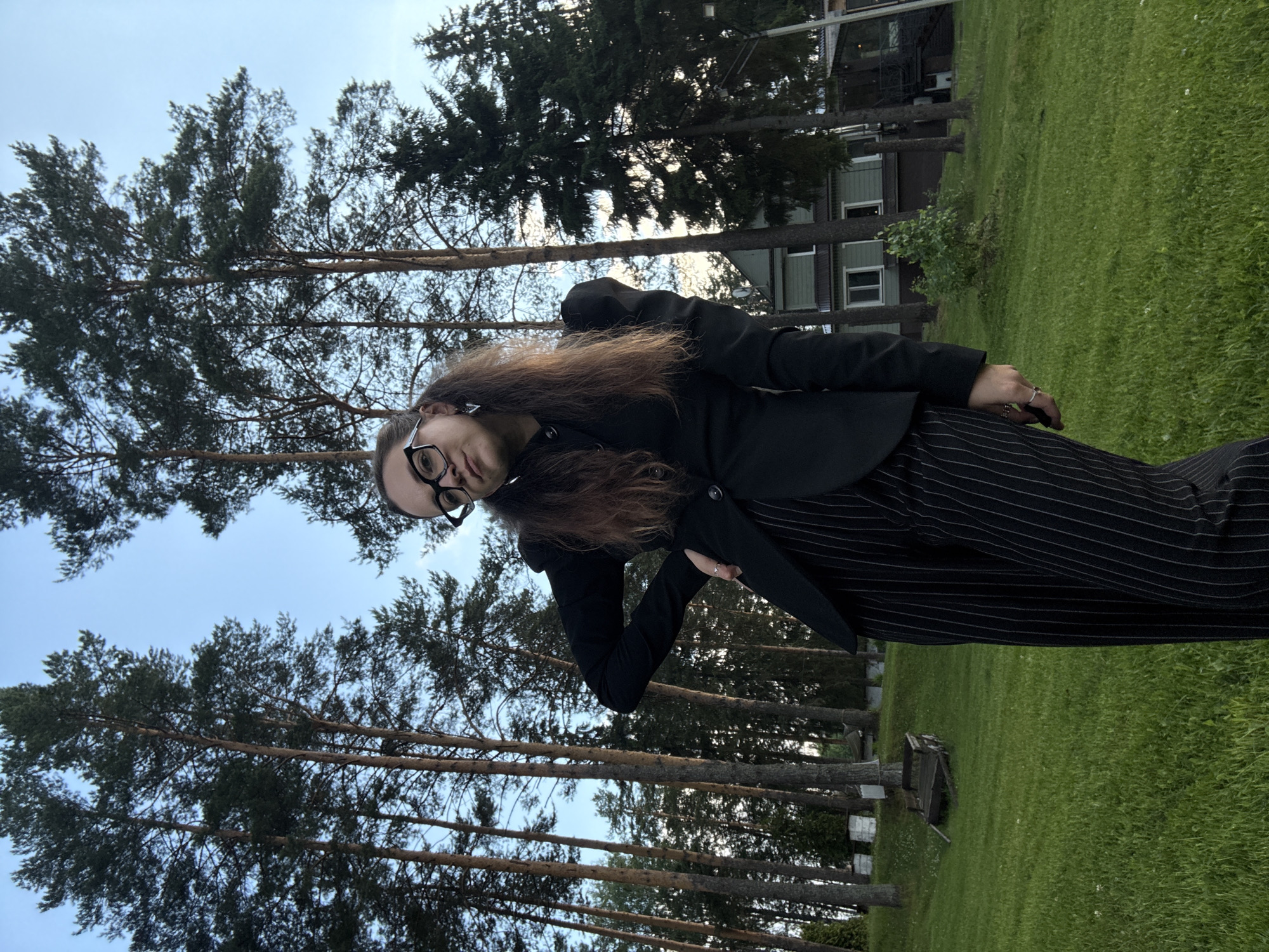 A young woman with long, brown hair styled in pigtails and wearing glasses stands in a grassy area with tall pine trees behind her. She is dressed in a black blazer with puffed shoulders and black pinstripe pants. Her left hand is on her hip. In the background, a light green two-story house with white trim is visible amongst the trees. The sky is a clear blue with a hint of overcast. The location is in the city of Votkinsk, Russia.