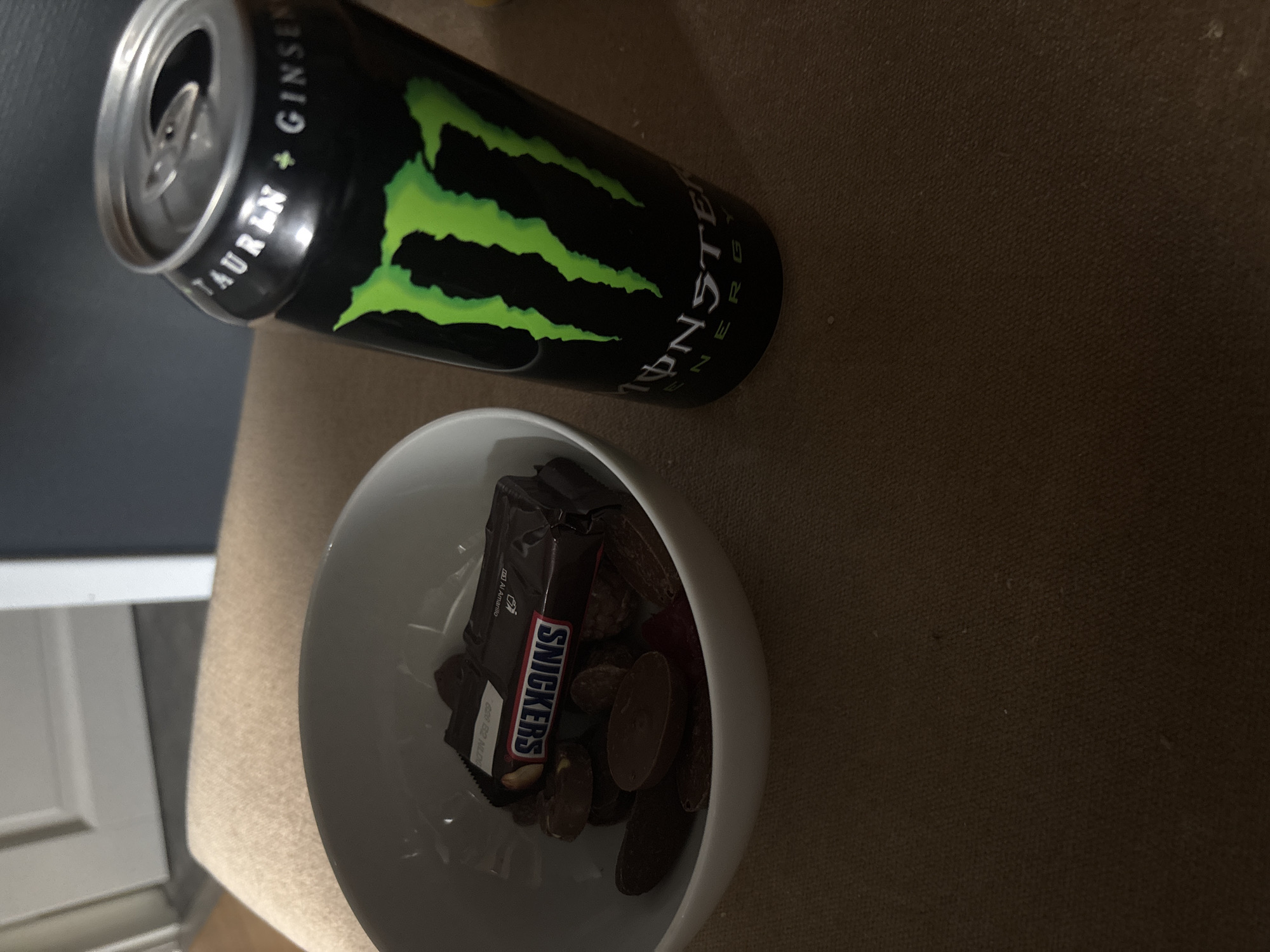 The image shows a close-up of a white bowl filled with chocolates and a single Snickers bar, next to a black Monster Energy drink can. The bowl is placed on a brown textured surface, likely a piece of furniture. The background is slightly blurred, showing a hint of a doorway and a wall. The scene is indoors and appears to be a casual setting. There is no indication of the location being in Egersund, Norway.