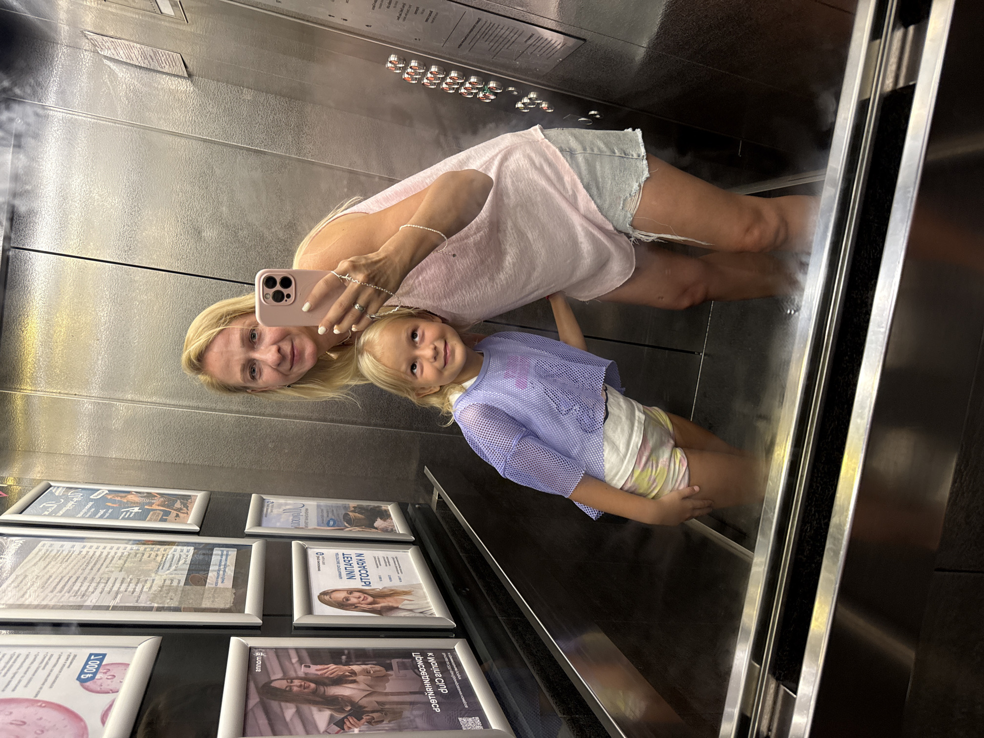 In the city of Yalta, Ukraine, a woman and a young girl are taking a selfie in an elevator. The woman, with blonde hair and fair skin, is wearing a light pink tank top and distressed denim shorts. She is holding a pink smartphone in her right hand, angled to capture the reflection. A young girl with blonde hair, wearing a sheer blue mesh top over a white top and tie-dye shorts, stands next to her, looking up and to the side. Both are reflected in the mirrored walls of the elevator. The elevator interior is stainless steel, with framed posters on the left wall. The floor of the elevator appears to be wet or steamy, with visible mist rising around their feet. The lighting is a mix of ambient light and reflection from the polished surfaces.
