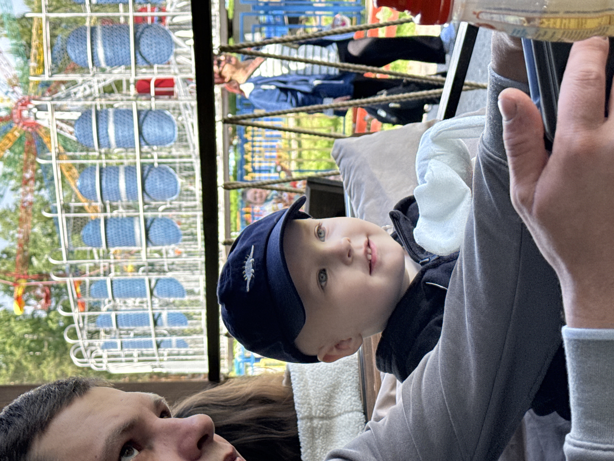 A close-up, canted-angle shot depicts a child with a blue cap looking upwards, seated with an adult in an outdoor setting. The location is identified as Chaikovsky, Russia, based on background elements.
The child is the primary subject, wearing a navy blue cap adorned with a white emblem. The child is wearing a black jacket with white fur interior. Their attention is directed upwards, slightly to the right.
The adult is positioned at the bottom left of the frame, partially cropped, with their face visible. Another person is partially visible behind the child.
In the background, a playground and a Ferris wheel with blue seats can be seen, establishing the location as an amusement park or similar recreational area. A person wearing a blue jacket is partially visible in the background.
The image appears to be taken from a low angle, emphasizing the child's upward gaze and the amusement park attractions. The lighting suggests it is daytime. A hand is visible at the top right of the frame, holding a cell phone.