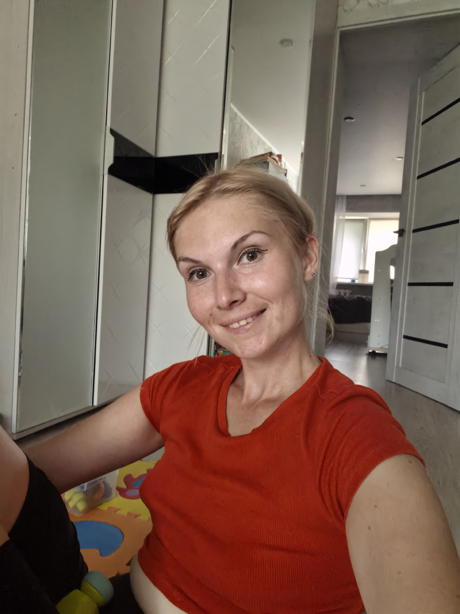 The image shows a woman with blonde hair and light-colored eyes smiling at the camera. She is wearing an orange t-shirt and black shorts. She is sitting on a colorful playmat with toys scattered around her. In the background, there is a closet with mirrored doors and a doorway leading to another room. The room appears to be a living space, possibly a child's room given the playmat. The lighting suggests it is daytime, and the interior setting indicates the photo was taken indoors. The woman's expression is cheerful and relaxed, suggesting a casual and comfortable moment. There is no visible text in the image. The location context of Sterlitamak, Russia, provides a general geographical setting.