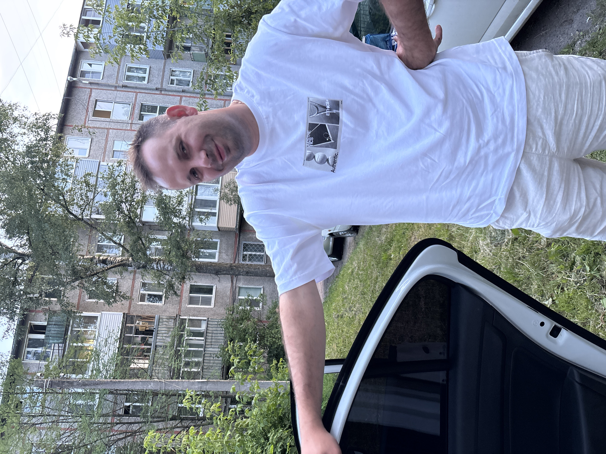 A man stands outside next to an open car door, leaning on the door frame. He is wearing a white t-shirt with a graphic print and light-colored shorts. Behind him is a multi-story apartment building, typical of Soviet-era architecture, with balconies and windows. Trees and greenery surround the building, and a patch of grass is visible on the ground. The scene appears to be in a residential area, possibly in Rusanovo, Russia, given the architectural style. The lighting suggests it is daytime.