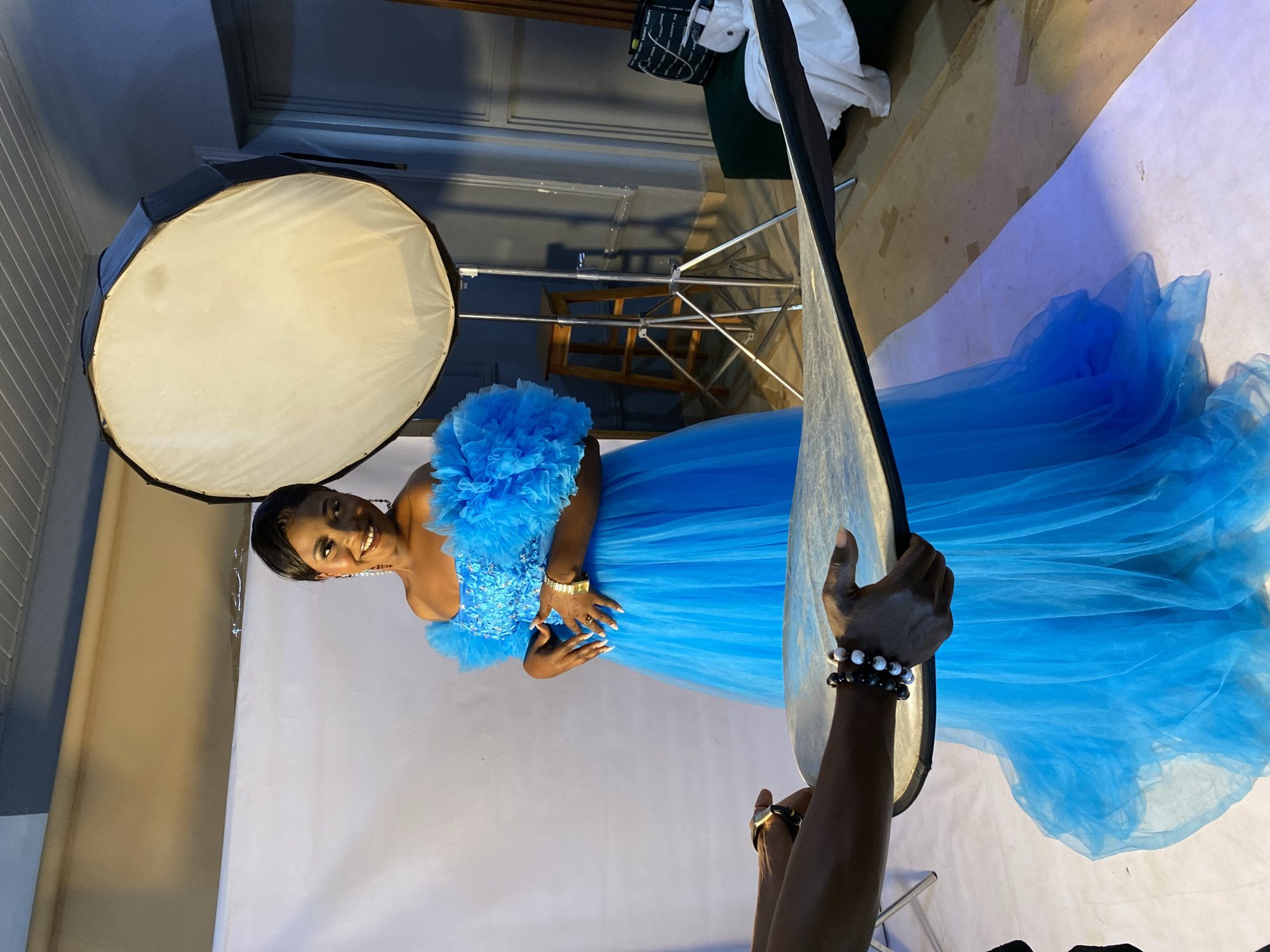 A woman in a vibrant blue, voluminous tulle gown poses in a studio setting. She has dark hair styled neatly and wears bright makeup, smiling warmly at the camera. Her gown features intricate beading on the bodice and a dramatic, ruffled off-the-shoulder design. The studio backdrop is a seamless white paper, with a large softbox light positioned to her left, illuminating the scene. A silver light stand and a reflector, held by another person whose hands are visible at the bottom right of the frame, are also present. The reflector has beaded bracelets on the wrist of the person holding it. The lighting suggests it is likely daytime, as it's a controlled studio environment. The overall mood is celebratory and glamorous. No visible text is present in the image.