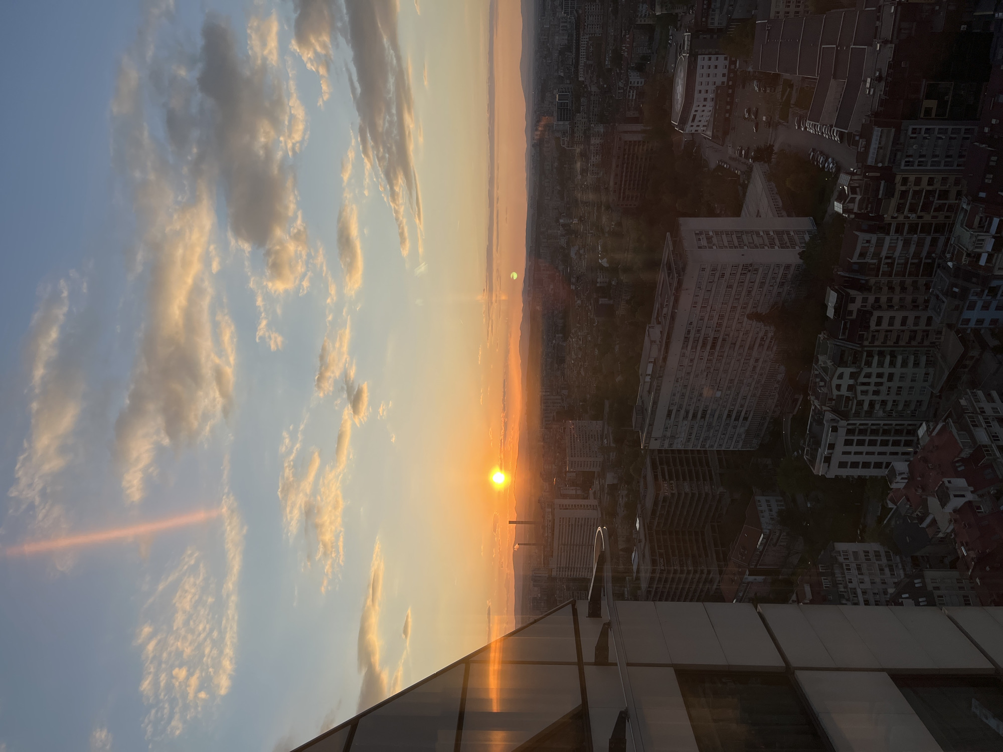 The image shows a sunset over Sofia, Bulgaria, as viewed from a high vantage point, possibly a tall building. The sun is low on the horizon, casting an orange glow across the sky, clouds, and cityscape. The sky on the left of the image is filled with clouds in varying shades of white and yellow, indicating the late afternoon or early evening.
To the right of the sunset is the cityscape of Sofia, with numerous buildings of varying heights and architectural styles. Some buildings are large, likely apartments or office complexes, while others are smaller residential structures. Streets and roads can be seen weaving through the buildings, creating a network of urban infrastructure.
The foreground features the structure of the building from which the photo was taken, with portions of the building visible on the bottom and right side of the image. The sun's light is reflected in the windows of the building.
