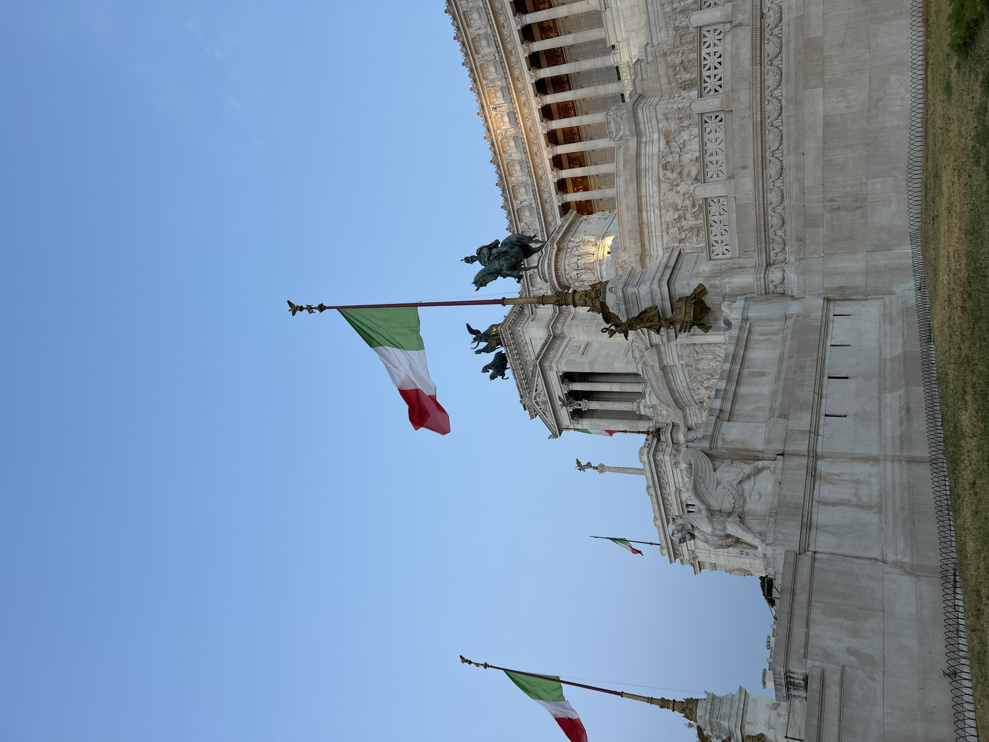The image captures a low-angle view of the Altare della Patria, also known as the Monument to Victor Emmanuel II, in Rome, Italy. The monument, constructed of white marble, dominates the scene. Several Italian flags are prominently displayed on flagpoles. The flag in the foreground, slightly to the left of center, is fully unfurled, showcasing its green, white, and red vertical stripes. Another Italian flag is visible in the upper right quadrant of the image, partially unfurled.
The monument itself features classical architectural elements, including columns and intricate sculptures. A large equestrian statue is visible atop a section of the monument, and other statues and reliefs adorn its facade. The sky above is a clear, pale blue, indicating it is either early morning or late evening. In the lower portion of the image, a patch of dry grass is bordered by a black metal fence. No people are visible in the frame.